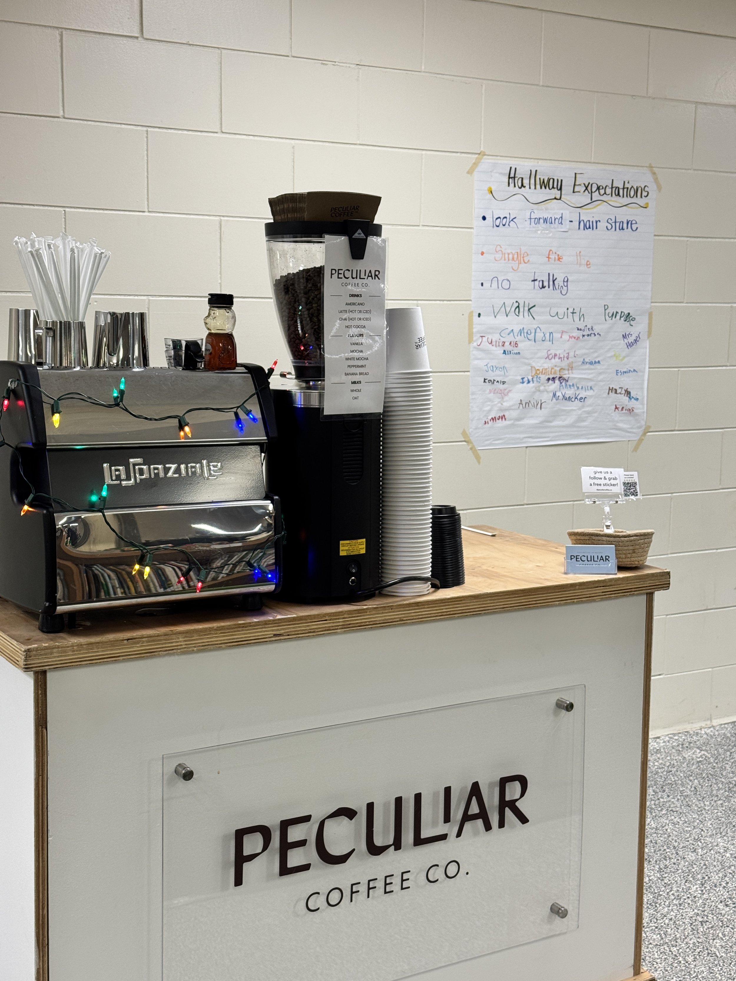 A coffee cart with a Peculiar Coffee Co. sign on a white counter, decorated with colorful string lights, and a white brick wall in the background.