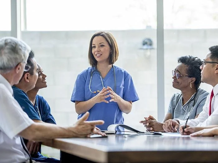 A diverse group of medical professionals, including a nurse in blue scrubs with a stethoscope, gathered around a table with a female nurse in blue scrubs standing and speaking during a meeting or discussion.