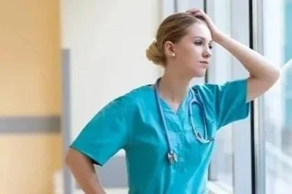 Nurse in blue scrubs looking out a hospital window with a contemplative expression.