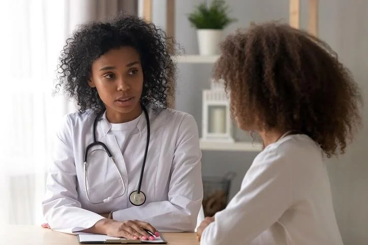 Two women, both wearing white coats and one with a stethoscope around her neck, are engaged in a conversation in a well-lit medical or clinical setting. One woman appears to be a healthcare professional, listening attentively to the other.