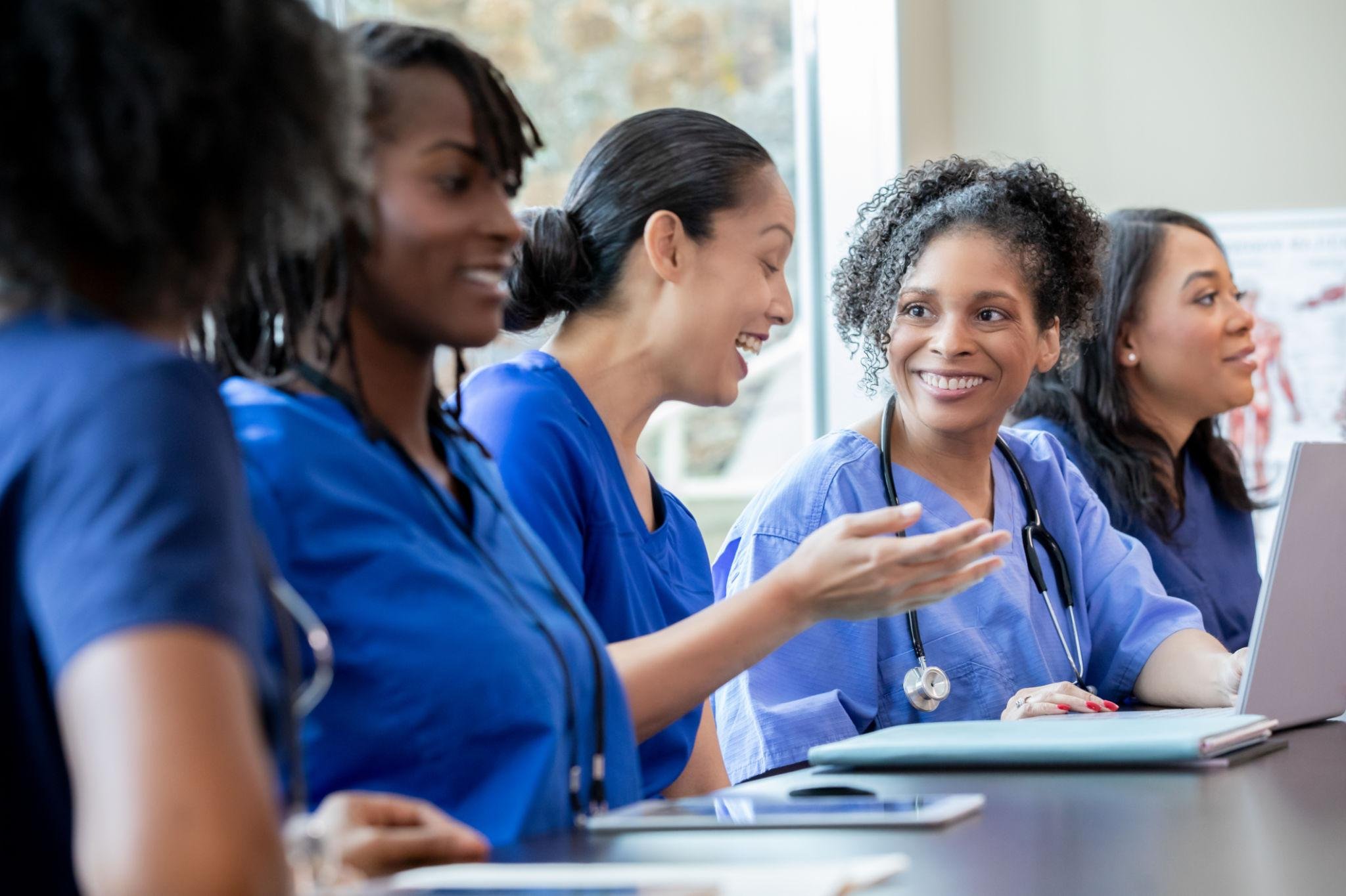 Group of diverse female healthcare professionals sitting at a table, engaging in discussion, wearing blue scrubs, some with stethoscopes, in a well-lit room with a window in the background.