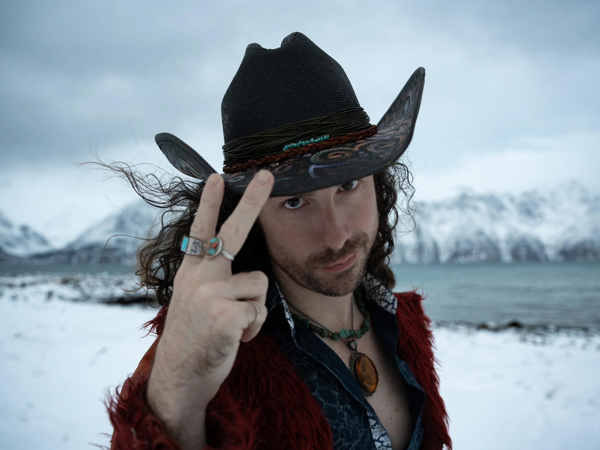 Man with curly hair and beard wearing a cowboy hat, rings, and a necklace, standing outdoors in a snowy landscape with mountains in the background.