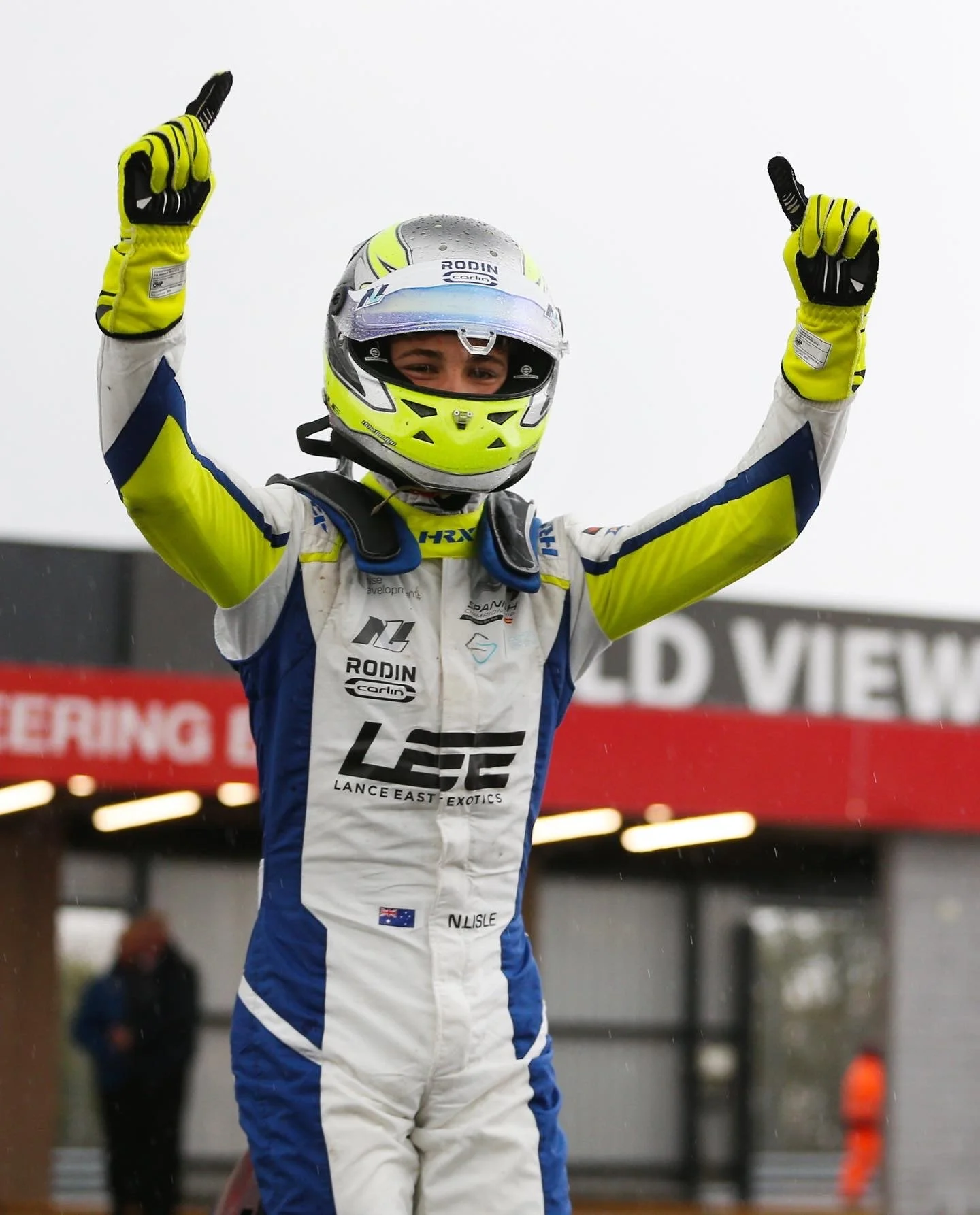 A young race car driver in a racing suit and helmet celebrating with raised arms at a racing event.