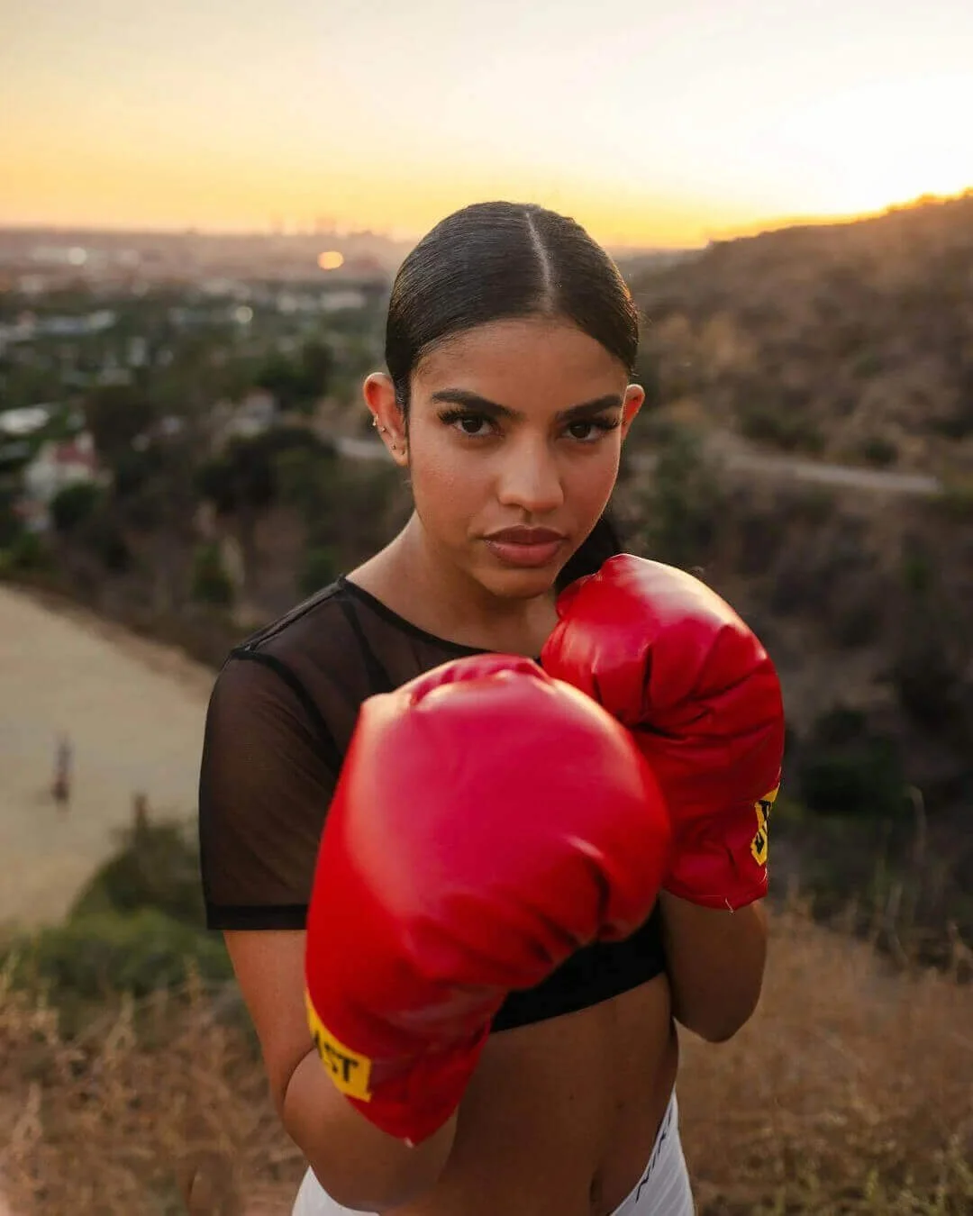 A young woman in boxing gloves poses outdoors during sunset, standing on a hill with a view of a city in the background.