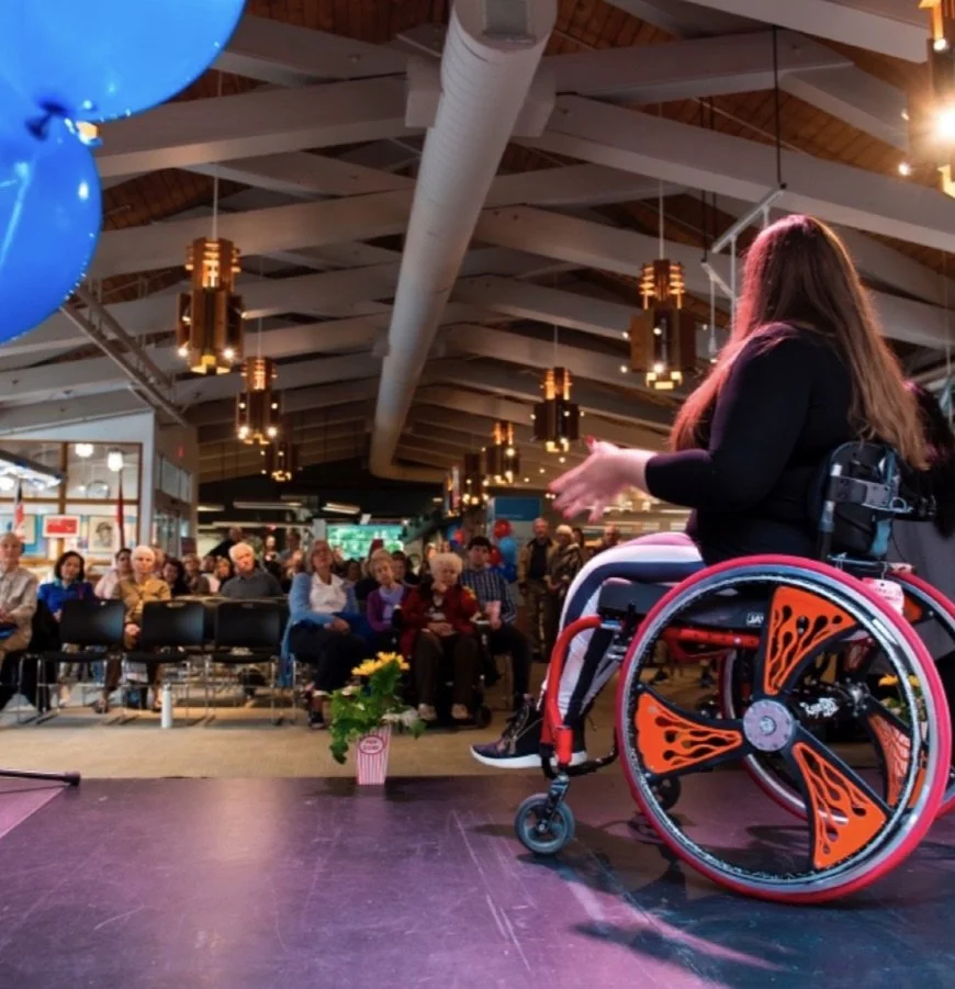 Jenna Reed-Côté using a wheelchair speaks to an audience from a stage in a large indoor community space. Viewed from behind and slightly to the side, addressing a seated crowd listening attentively. Her wheelchair features bright orange flame spoke