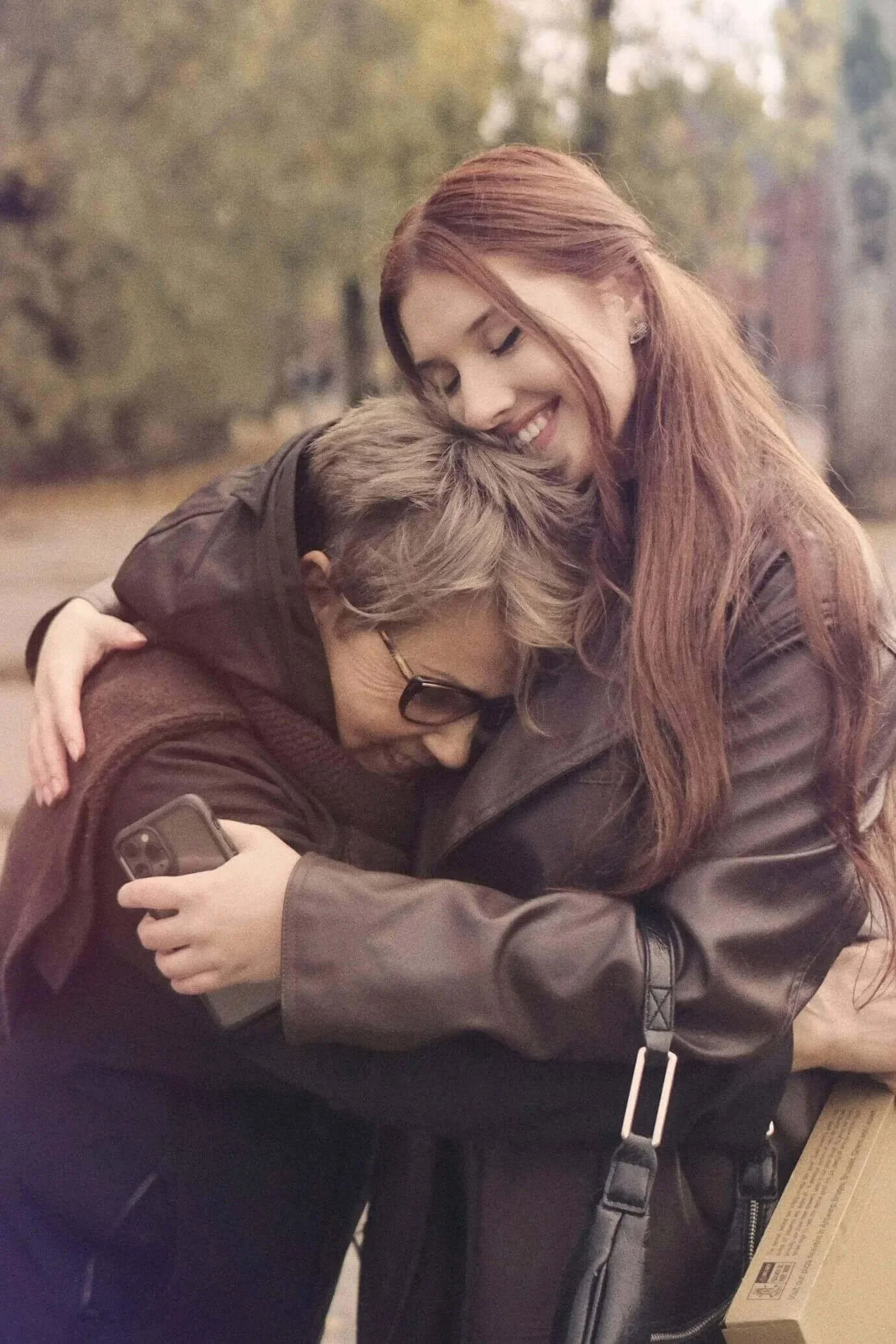 Two women hugging outdoors, one older and one younger, both smiling and holding a phone, with trees in the background.