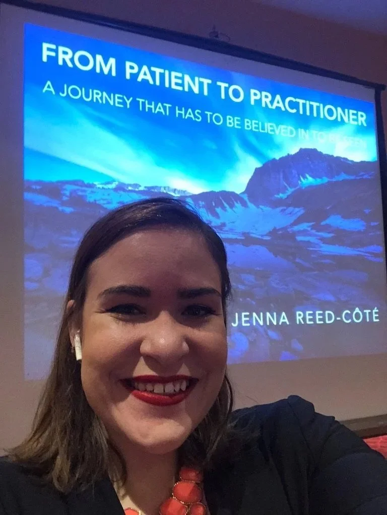 Jenna Reed-Cote smiling in front of a projected presentation slide titled "FROM PATIENT TO PRACTITIONER: A JOURNEY THAT HAS TO BE BELIEVED IN TO BE SEEN." She is wearing a dark blazer, red lipstick, a coral statement necklace and a wireless earbud.