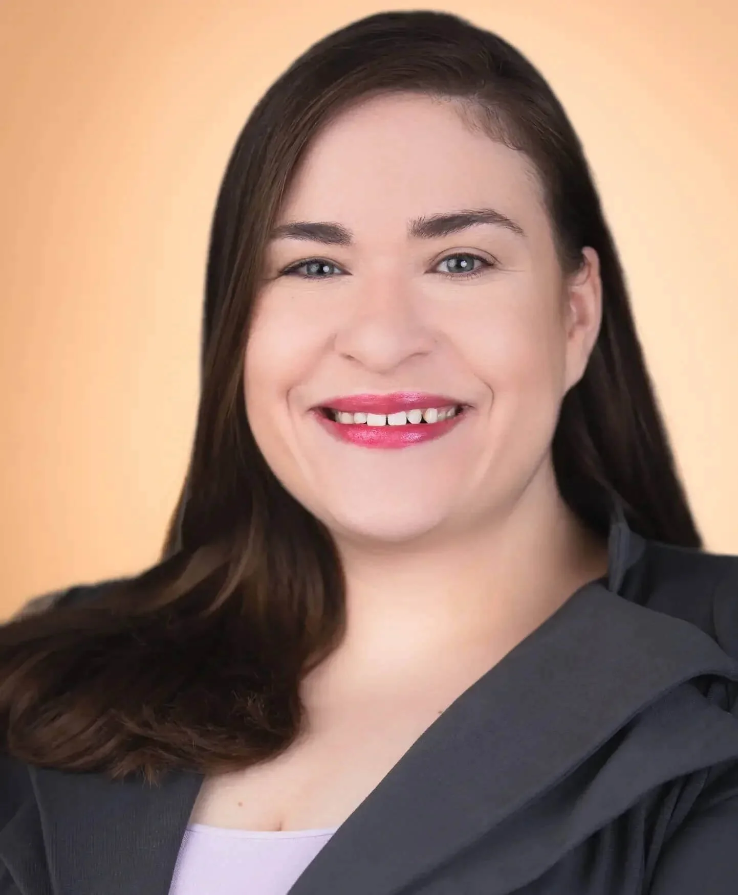 A woman with long brown hair, wearing a black blazer and pink lipstick, smiling against a blurred, peach-colored background.