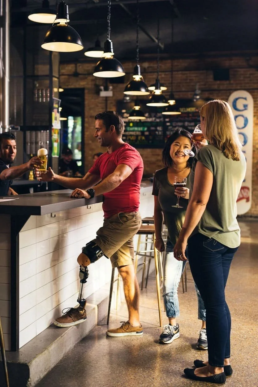 Three women and a man at a bar. The man, with a prosthetic leg, is receiving a beer from a bartender. The two women are holding drinks and smiling, engaging in conversation in a modern, dimly lit bar with hanging pendant lights and a brick wall.
