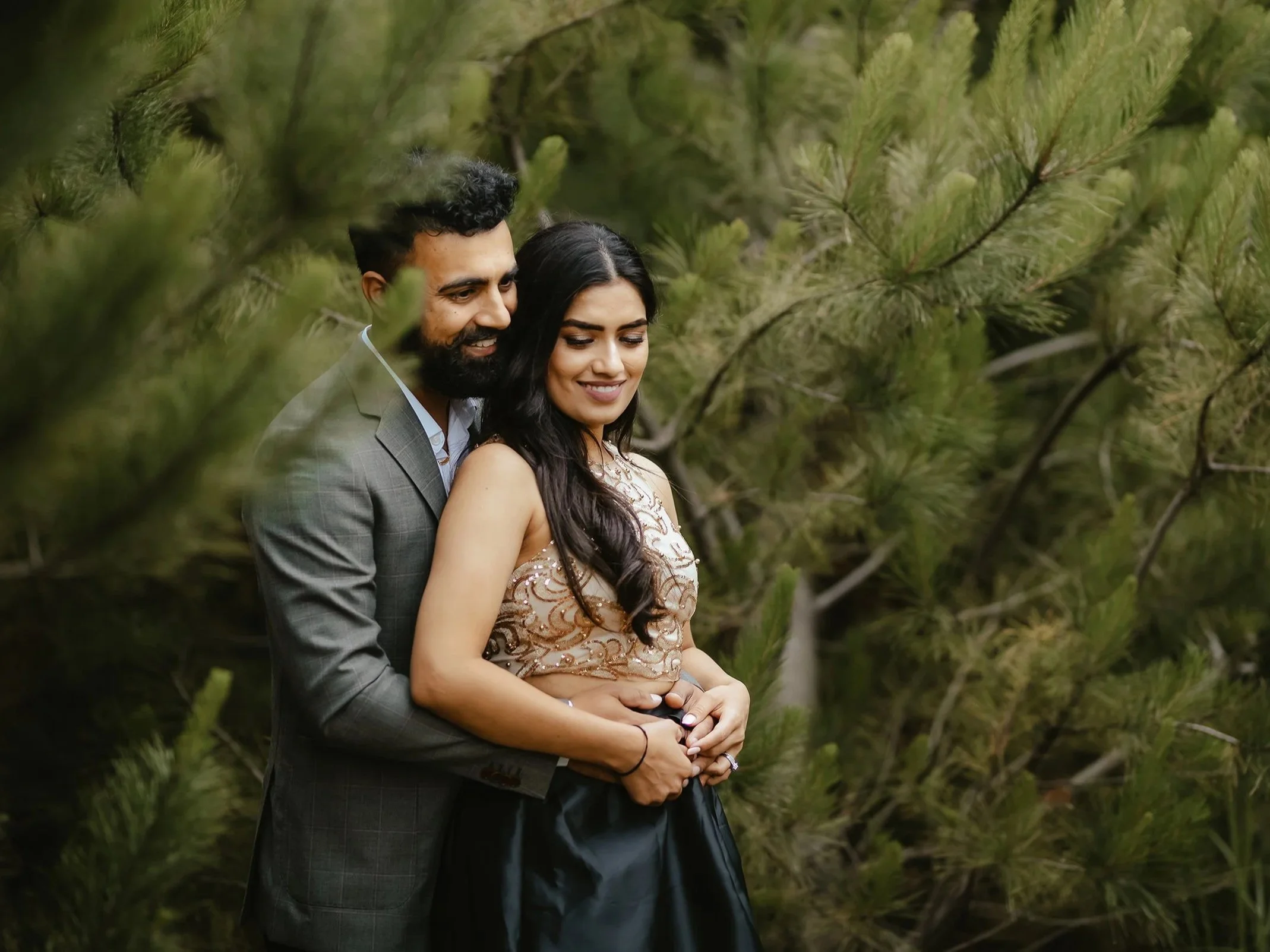 A couple is embracing outdoors among green pine trees, smiling and enjoying each other's company.