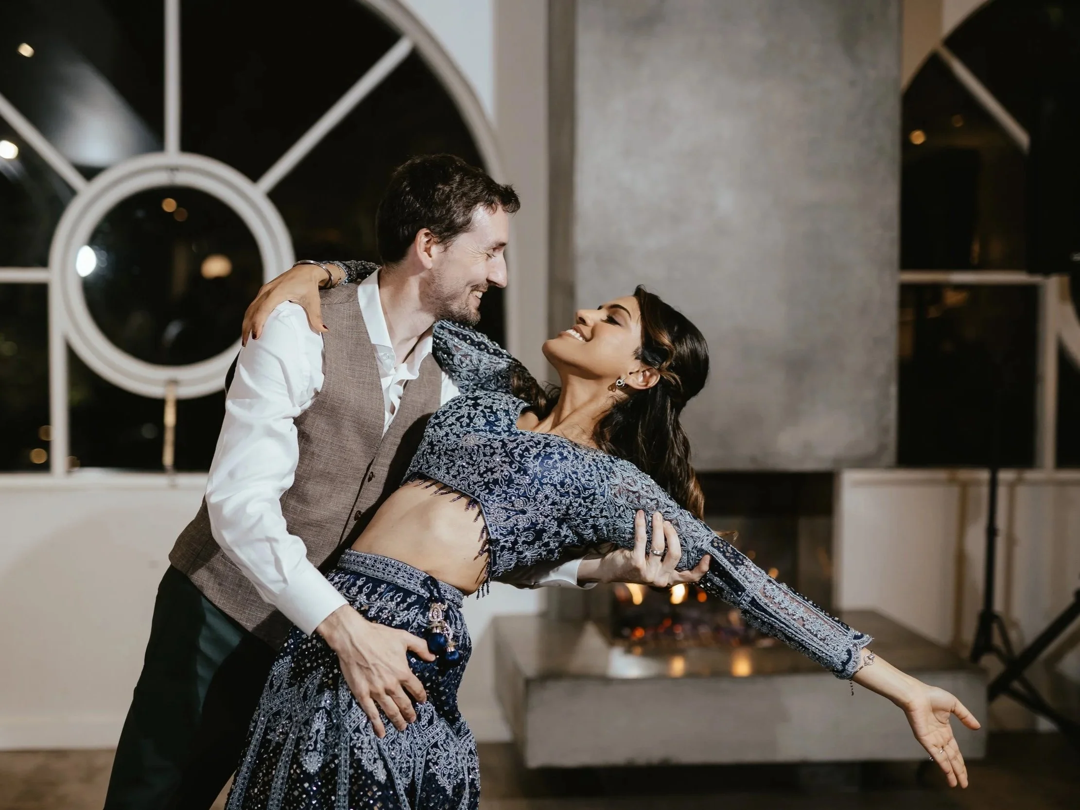 A man and woman dancing closely in a warmly lit room with large arched windows, with the man smiling and the woman reaching out with one arm, both dressed in elegant clothing.