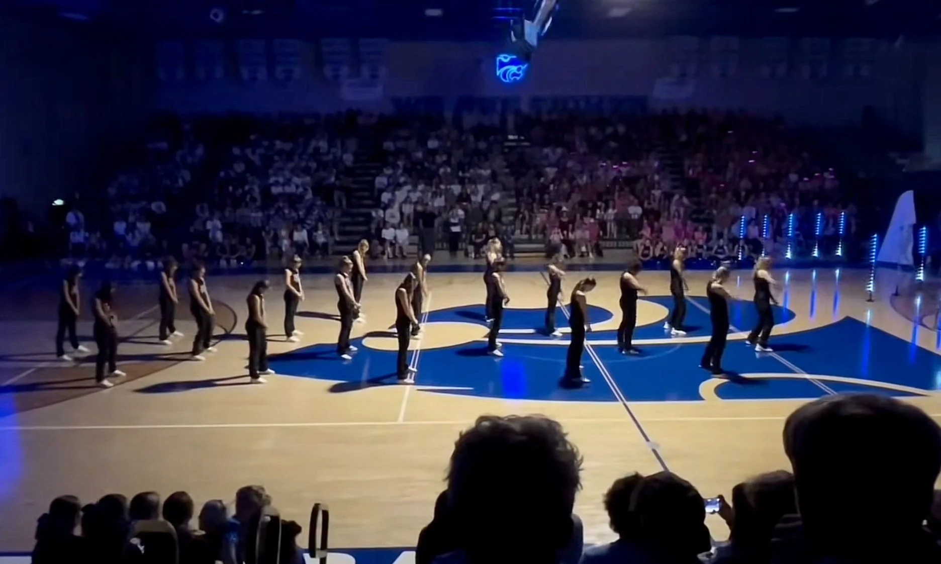 A group of dancers performing a routine on a basketball court, with audience members seated in the background.