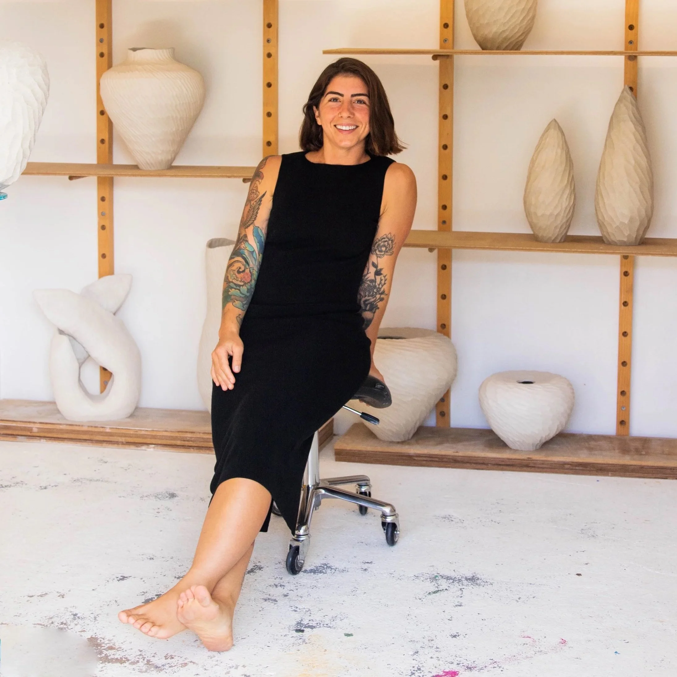 Smiling woman with dark hair, tattoos on arms, sitting on a rolling chair in a modern room with wooden shelves and white decorative vases.