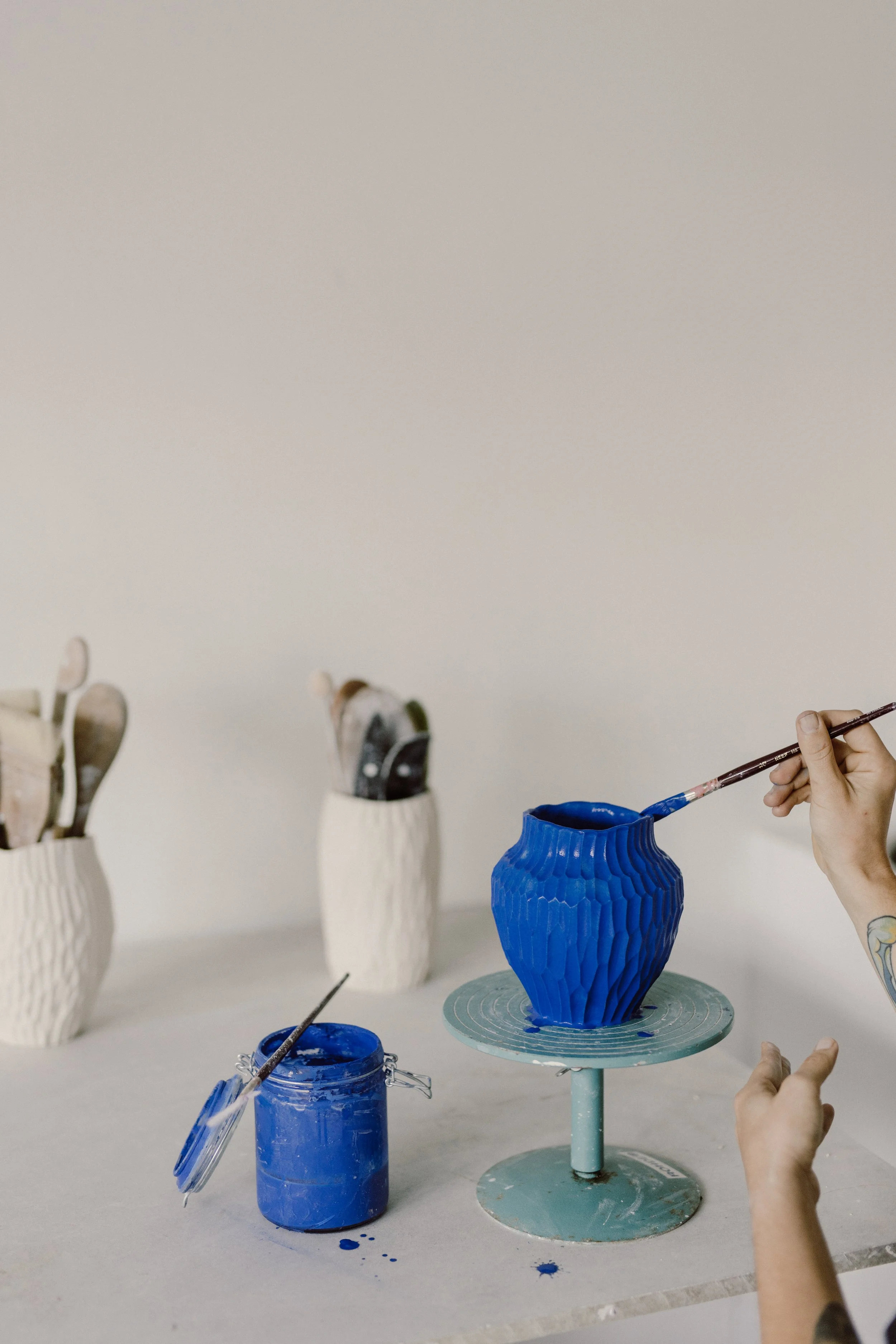 Person painting a blue ceramic vase at a pottery studio, with jars of blue paint and brushes on the table.