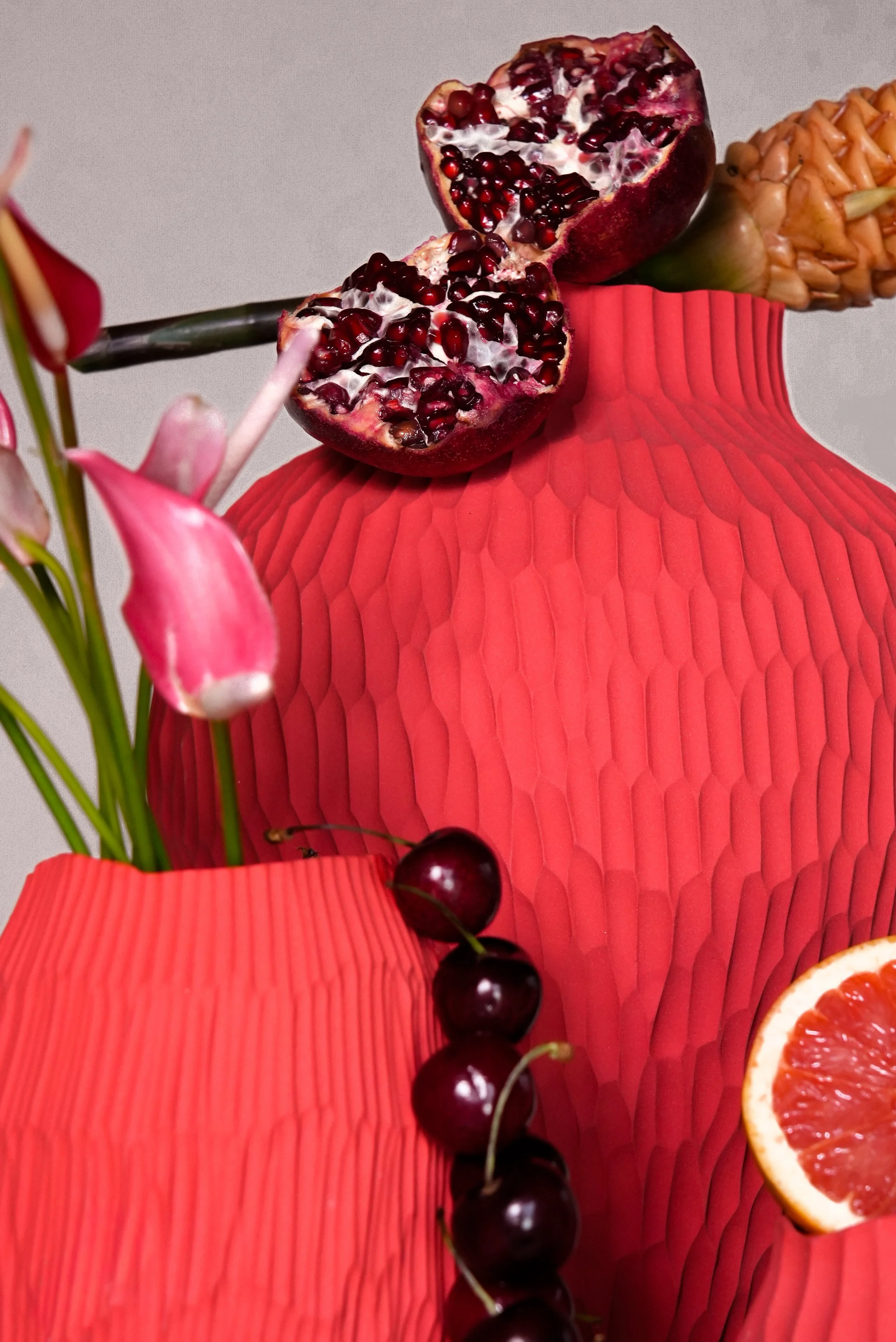 Red textured vases with pomegranate, cherries, pink lilies, and a grapefruit slice arranged on top.