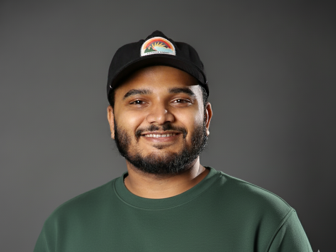 A man with a beard wearing a black cap and a green sweatshirt, smiling at the camera against a dark gray background.