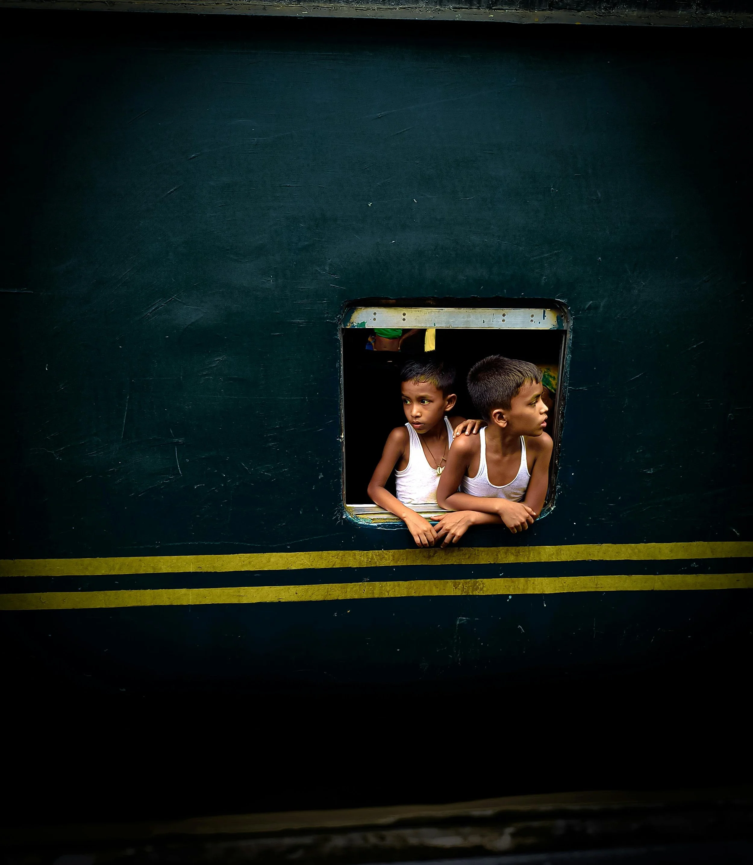 Two young boys in white tank tops look through a window on a dark green train.