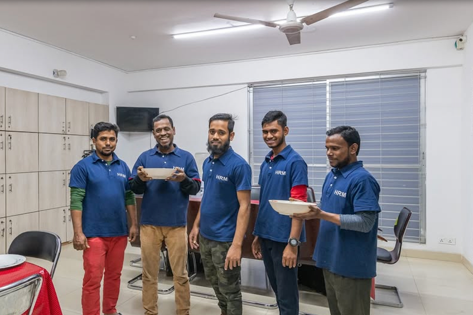 Five men standing in an office room, smiling. Four of them are holding plates and all are wearing blue shirts with 'HRM' logo. The room has lockers, a window with blinds, and a table with a red tablecloth in the foreground.