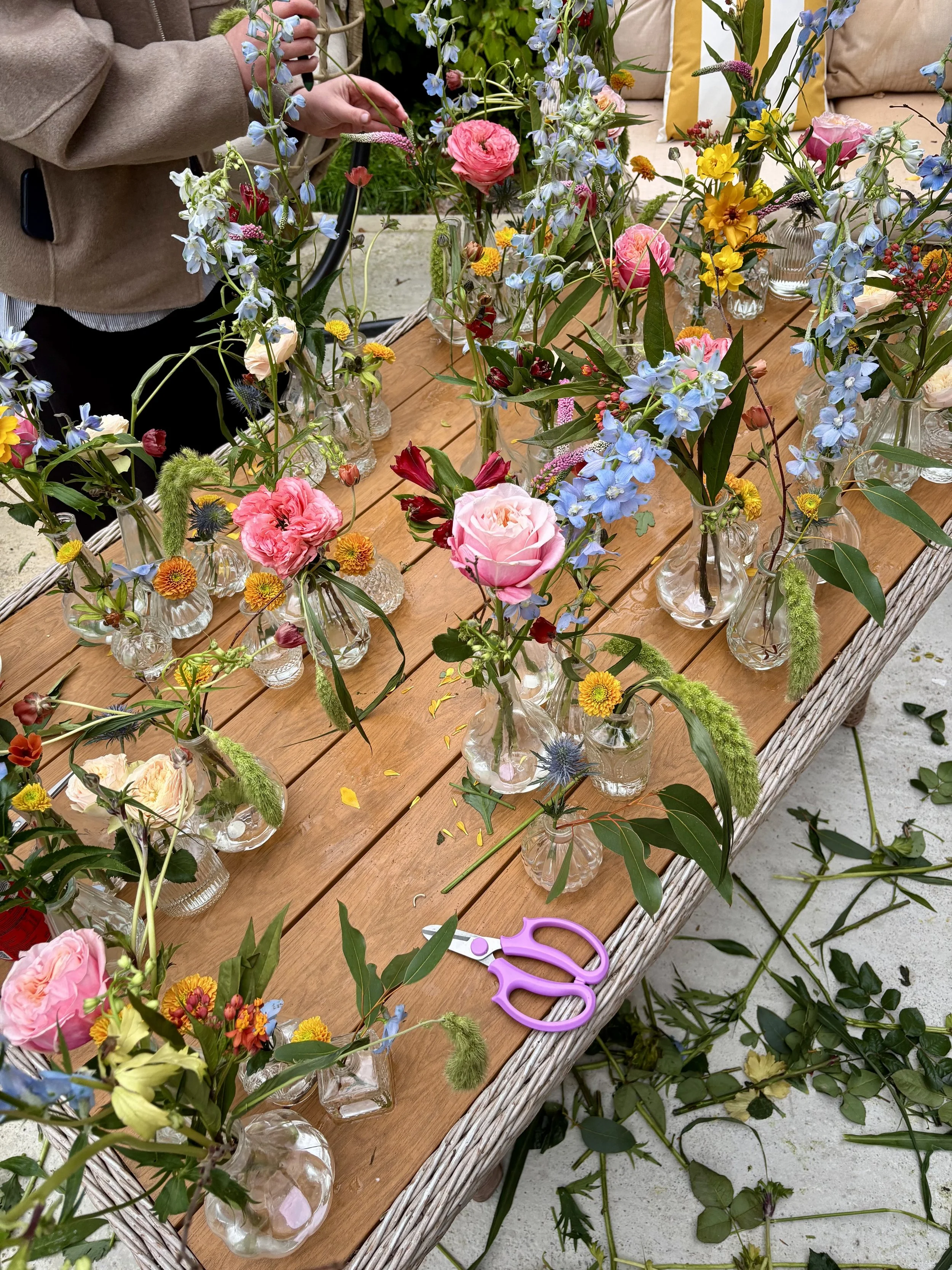A person arranging colorful flowers in small glass vases on a wooden outdoor table, with scissors nearby.