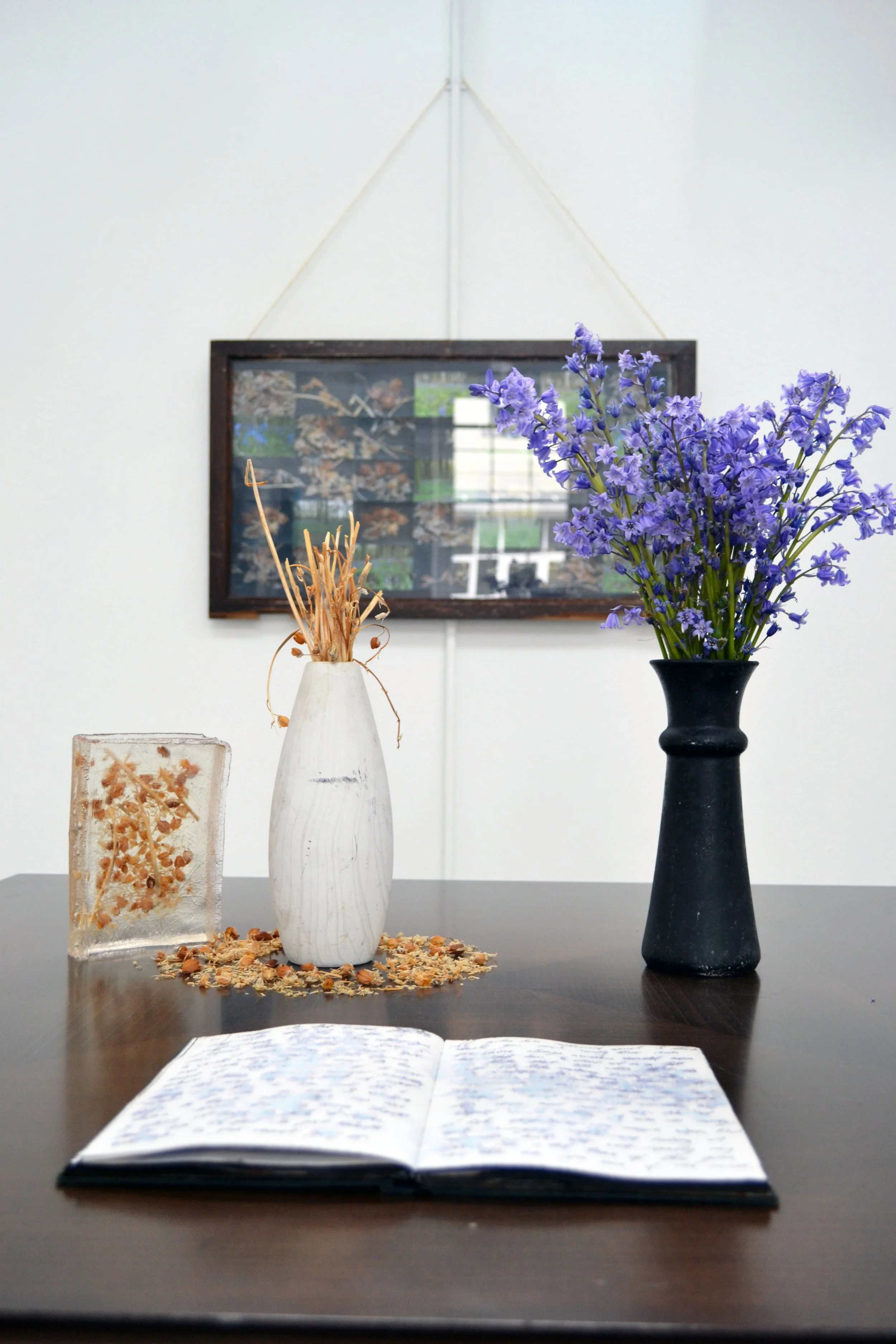 A dark wooden table with an open notebook, a white vase with dried flowers, a black vase with purple flowers, and a square glass block with dried flowers inside. In the background, a rectangular framed picture hangs on a white wall.