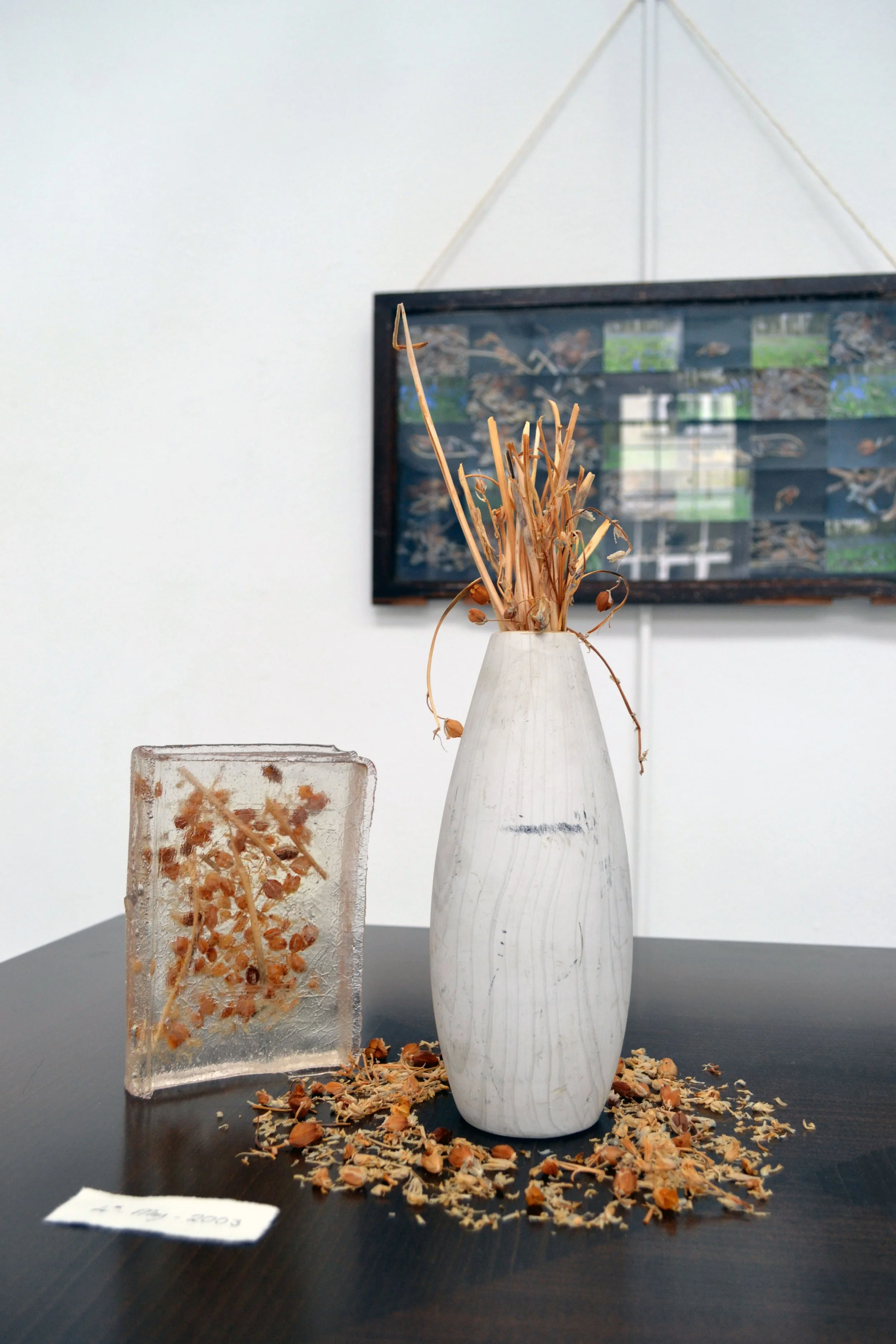 A white ceramic vase with dried flowers on a dark table, with a clear rectangular glass block containing more dried flowers beside it, and a framed collage of photographs on the wall in the background.