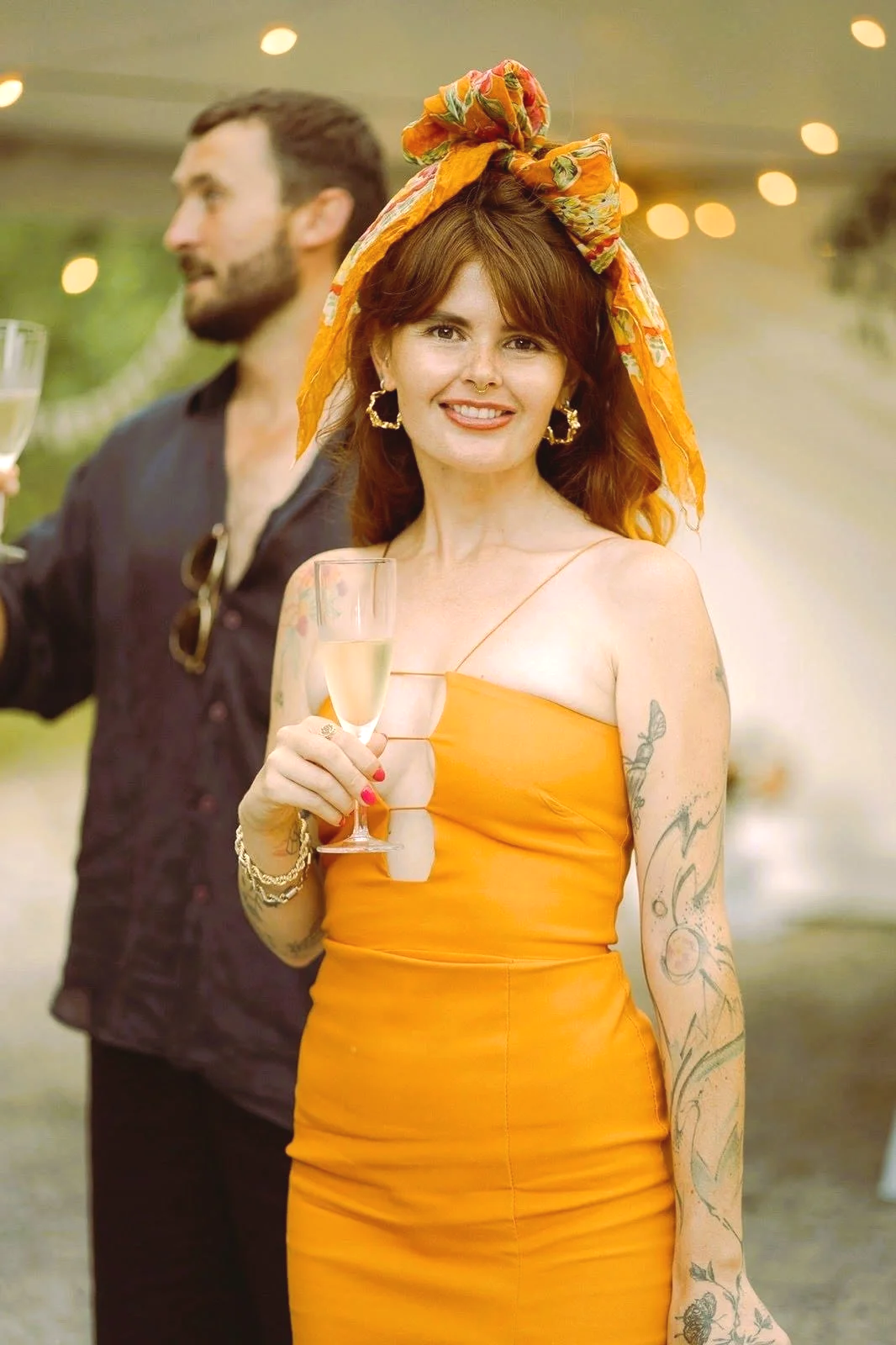A woman in an orange dress with tattoos, holding a glass of champagne at a celebration or outdoor event.