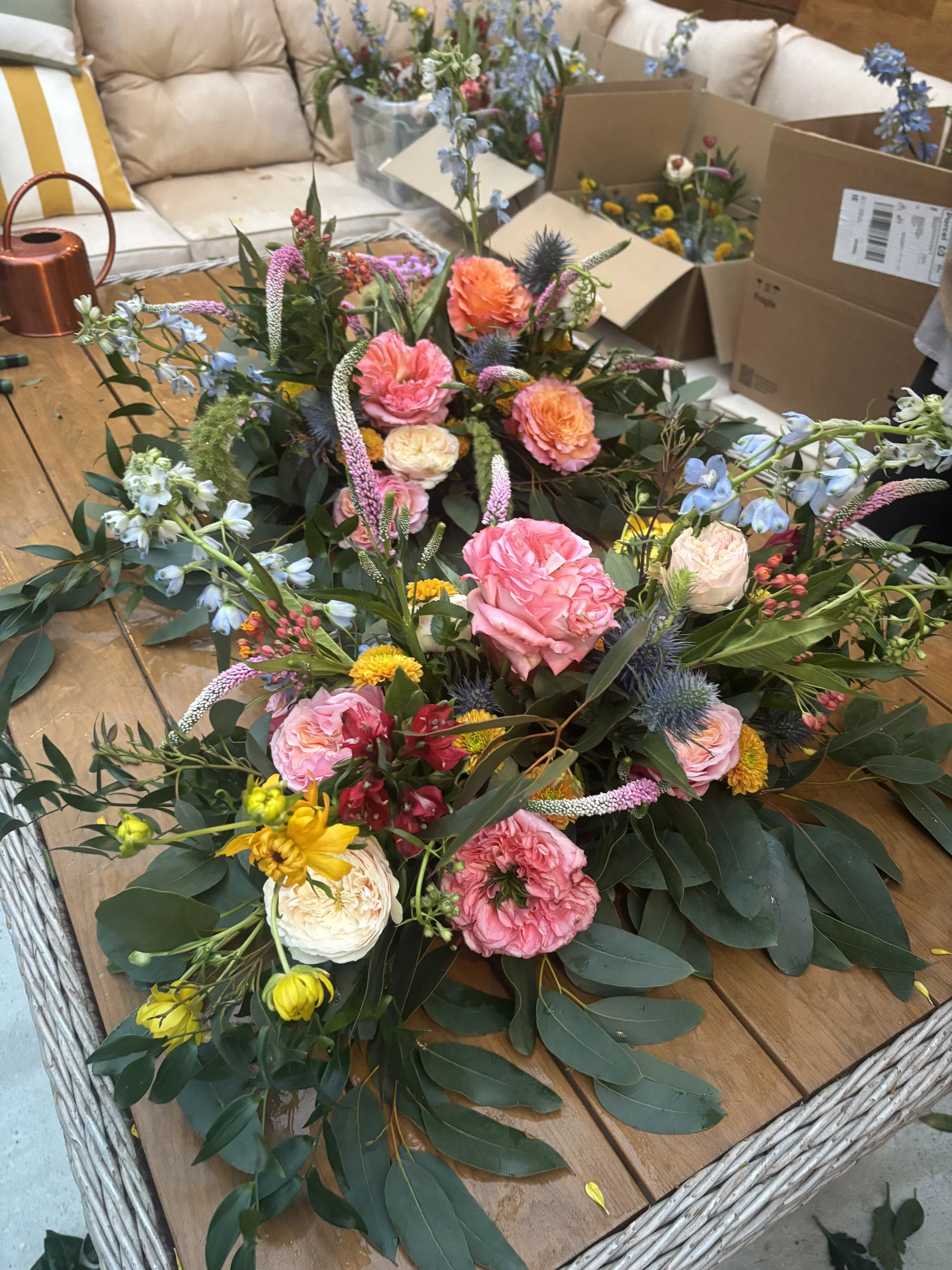 Assorted fresh flowers arranged on a wooden table with cardboard boxes in the background. The flowers include pink, peach, blue, yellow, and red blossoms with green leaves.