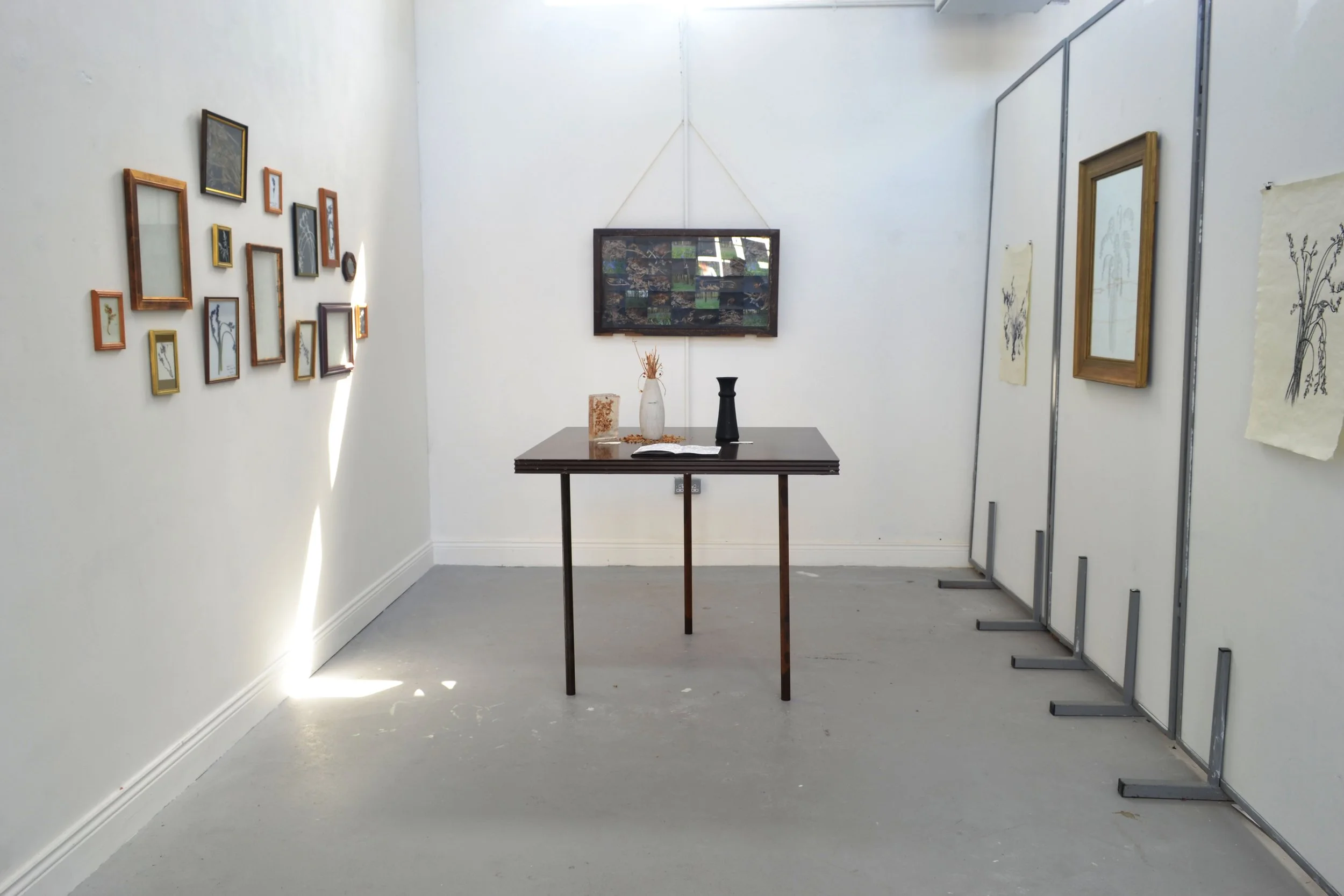 An art gallery with framed artwork on white walls, a wooden table with decorative items, and a foldable partition showing more art pieces.