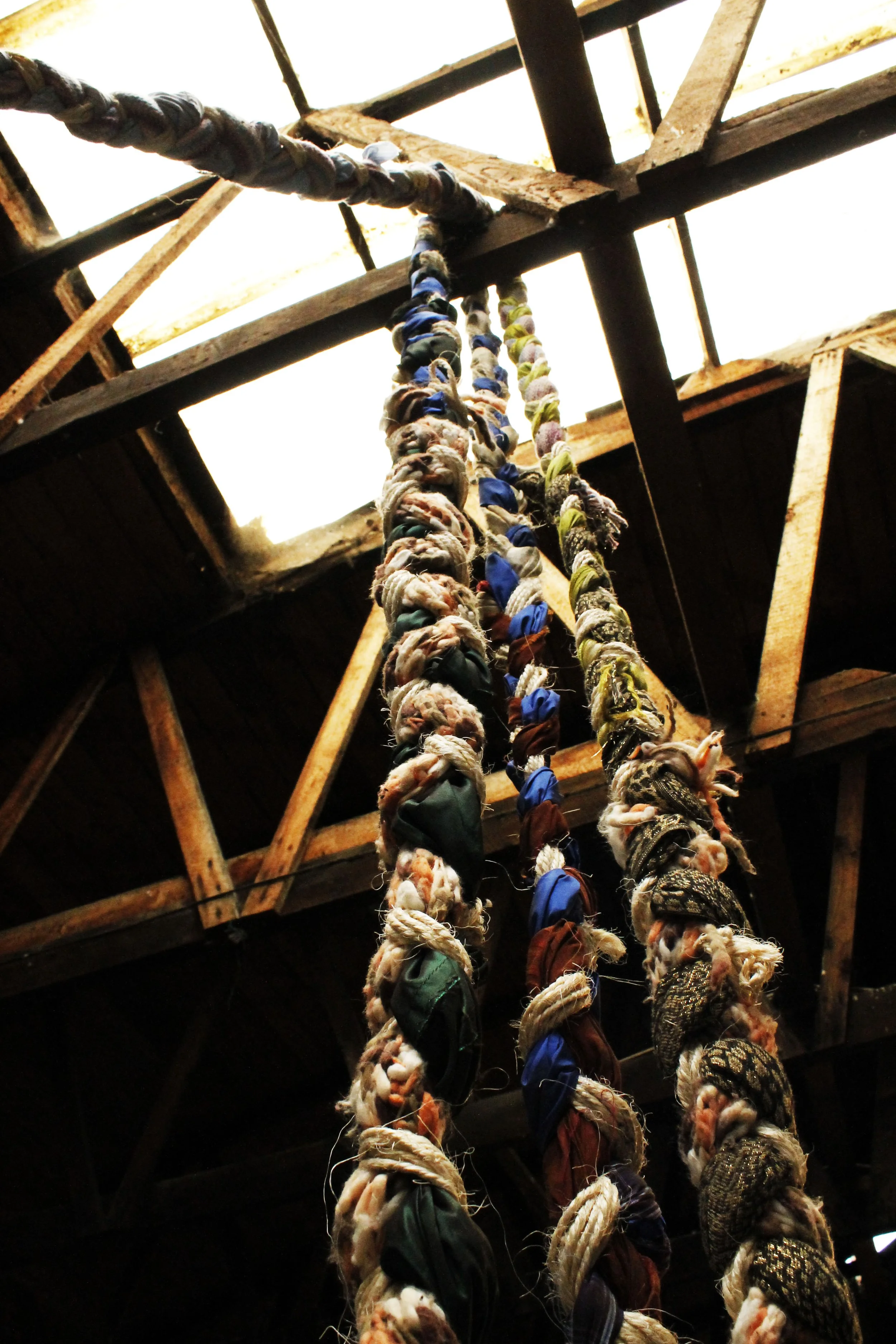 Colorful fabric rags tied to thick ropes hanging from a wooden ceiling with exposed beams.