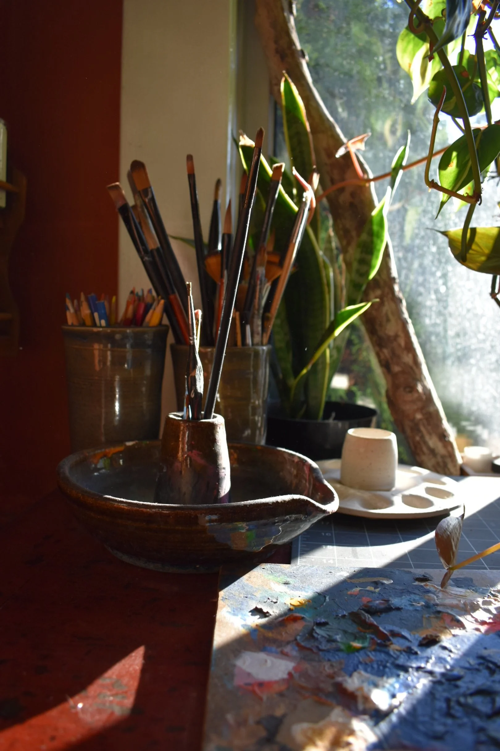 A sunlit artist's workspace with paintbrushes in a ceramic holder, potted plants, a palette with paint, and a window showing greenery outside.