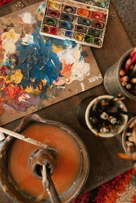 Paint palette with mixed colors, watercolor paints in a palette, and jars with colored pencils and brushes on a wooden table.