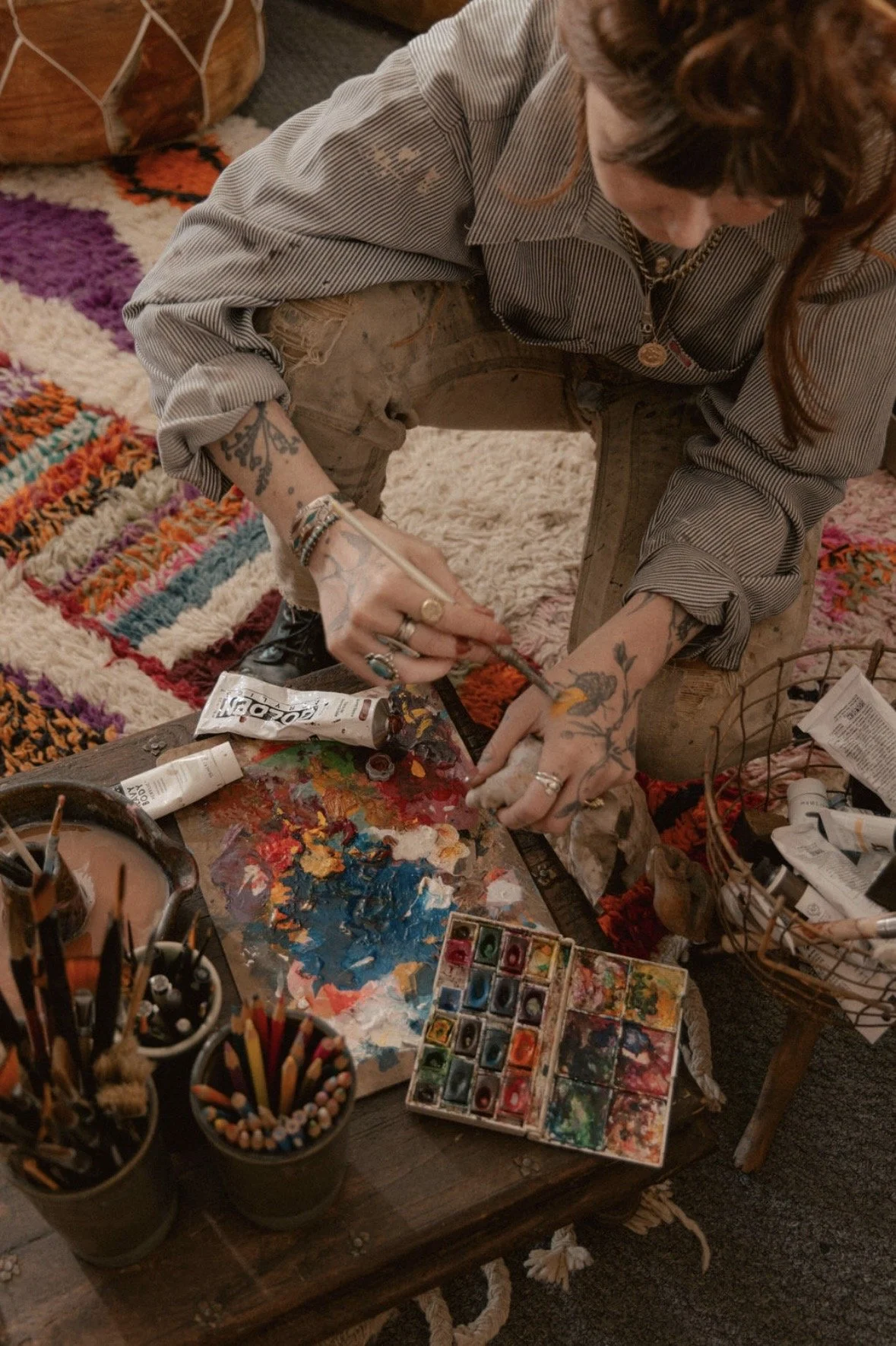 A person with tattoos, wearing a striped shirt with rolled-up sleeves, is painting with a small brush on a piece of fabric. The table in front has various art supplies, including paint tubes, a palette with colorful paints, and containers filled with brushes and pencils. The person has reddish-brown hair and is focused on their artwork, surrounded by a cozy indoor setting with a colorful rug, a basket, and other items.