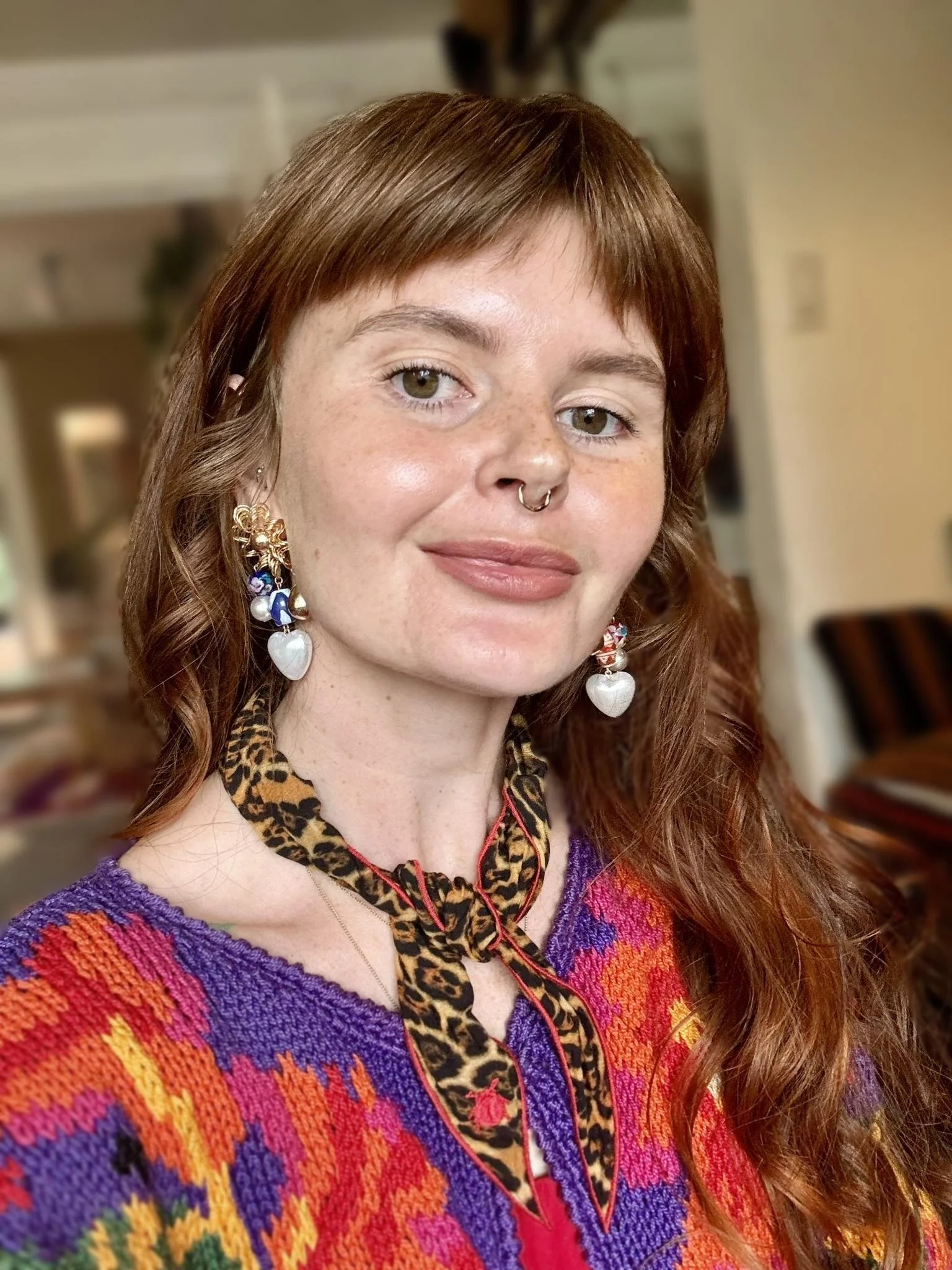 A woman with shoulder-length red hair, freckles, and a septum piercing. She is wearing colorful earrings, a leopard-print ribbon, and a vibrant multicolored sweater. The background shows an indoor setting with furniture.