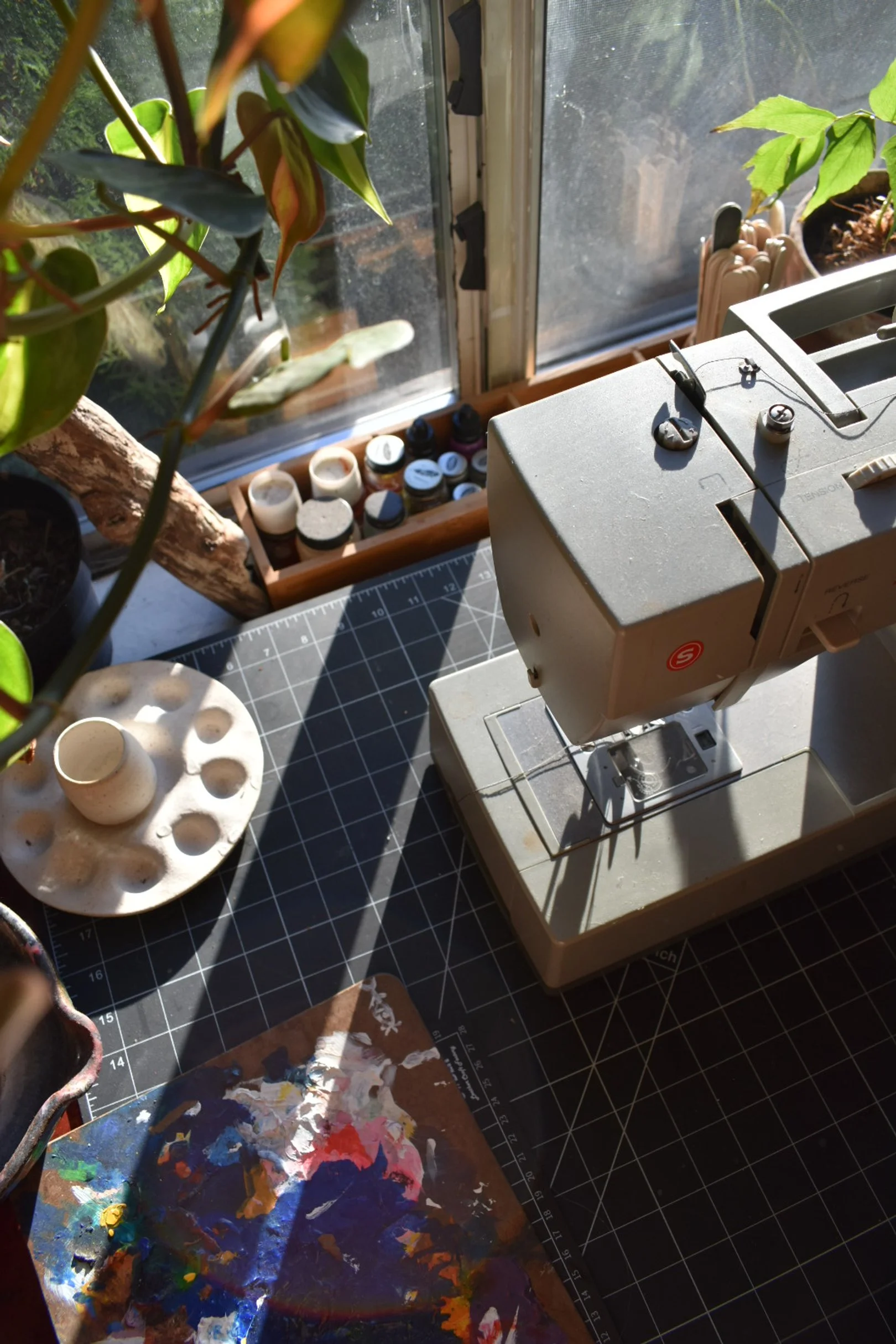 A sewing machine on a black cutting mat near a window with sunlight, surrounded by potted plants and art supplies.