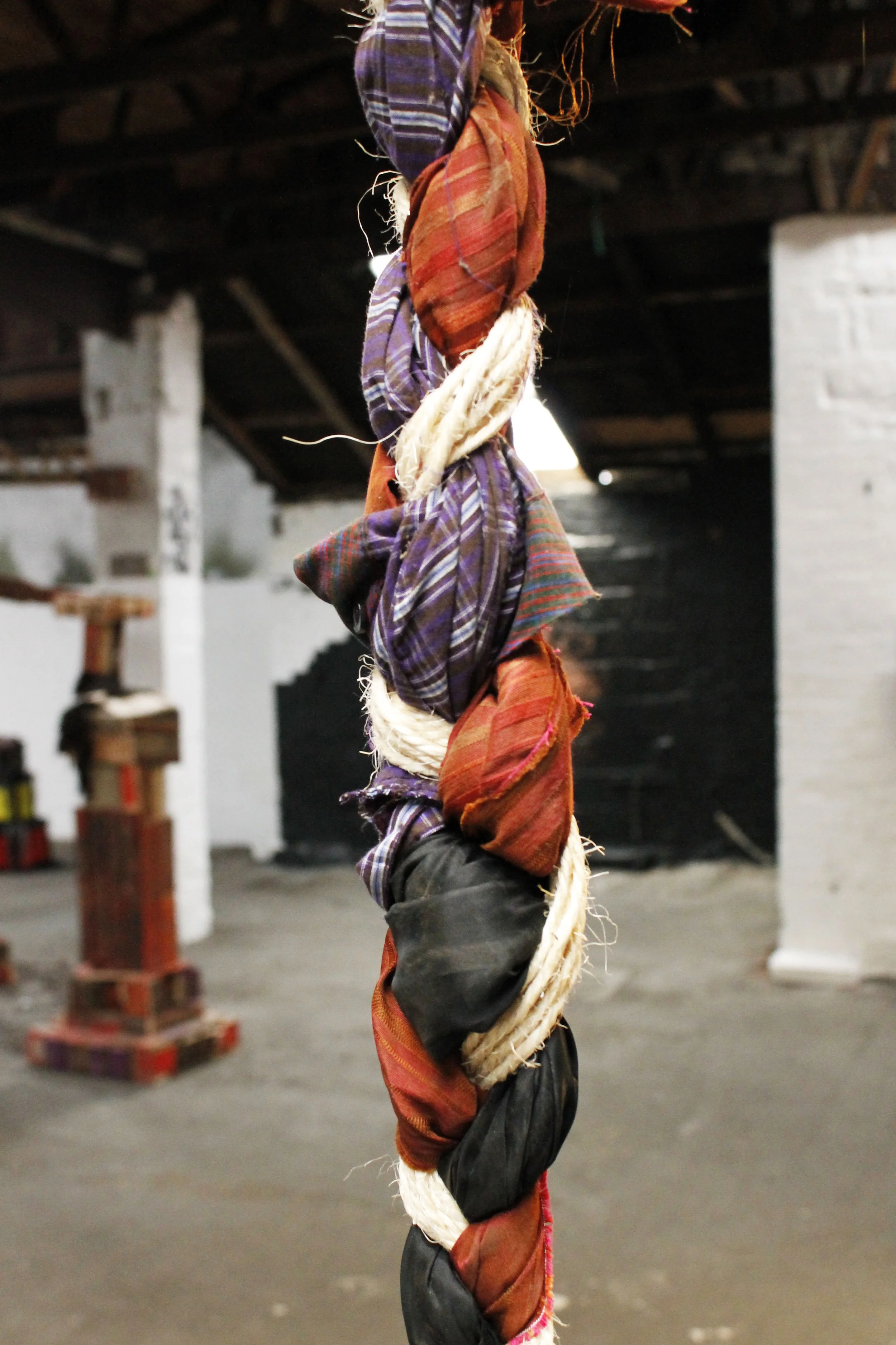 A twisted rope with fabric tied around it, featuring various colorful cloth pieces in purple, red, black, and cream, hanging vertically in an industrial or warehouse setting.
