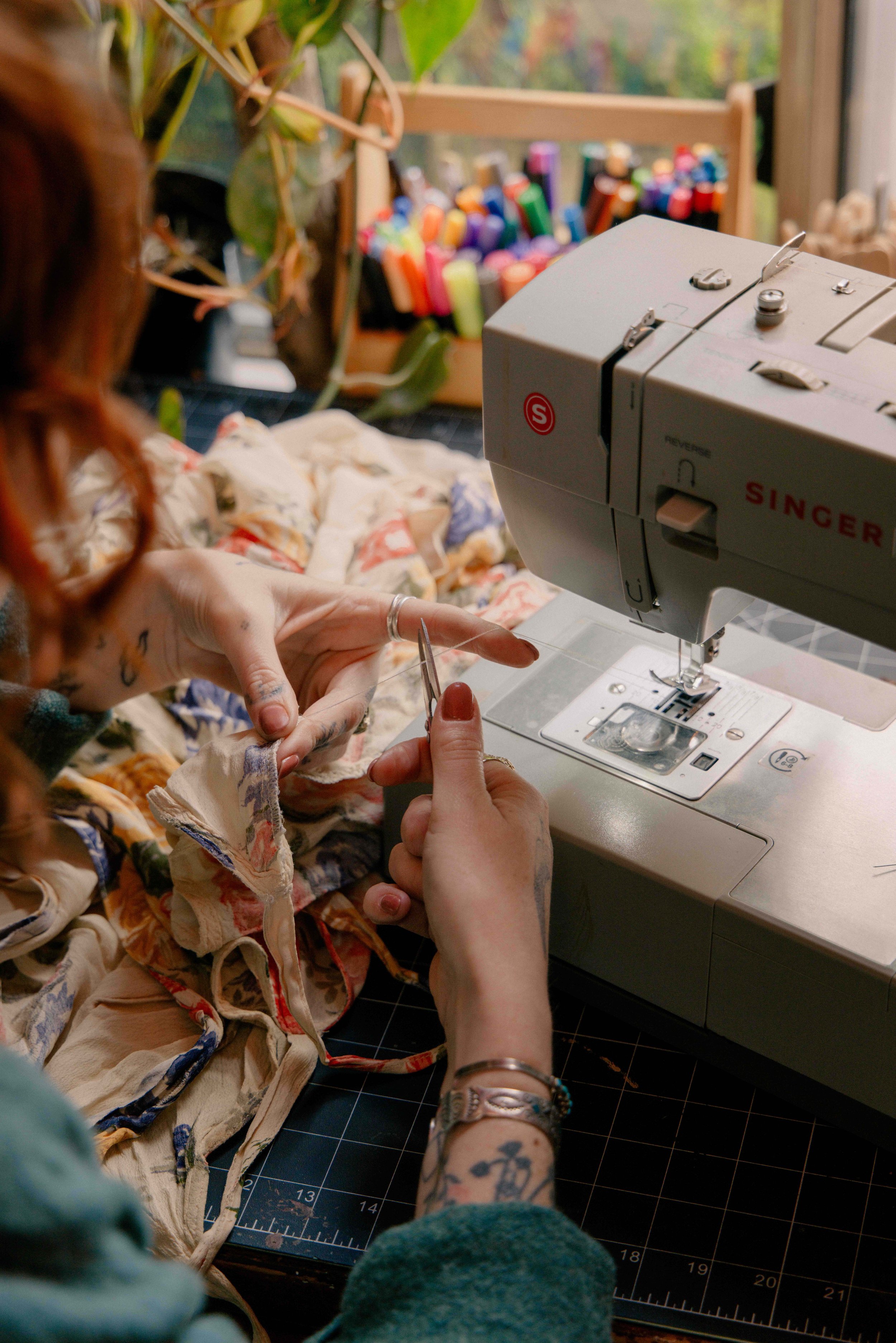 A person sewing fabric with a sewing machine, with colorful markers in the background and a person partially visible in the foreground.