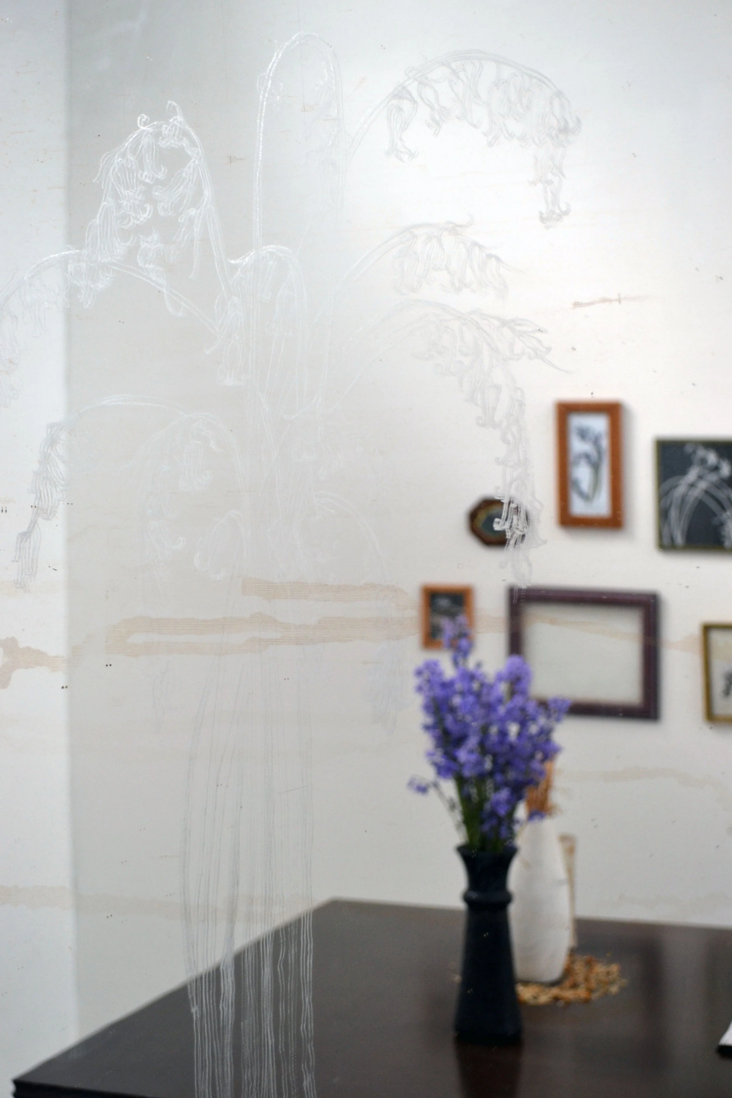 Reflection of a floral sketch on a glass wall, with a blurred interior scene including framed pictures and a vase of purple flowers on a table