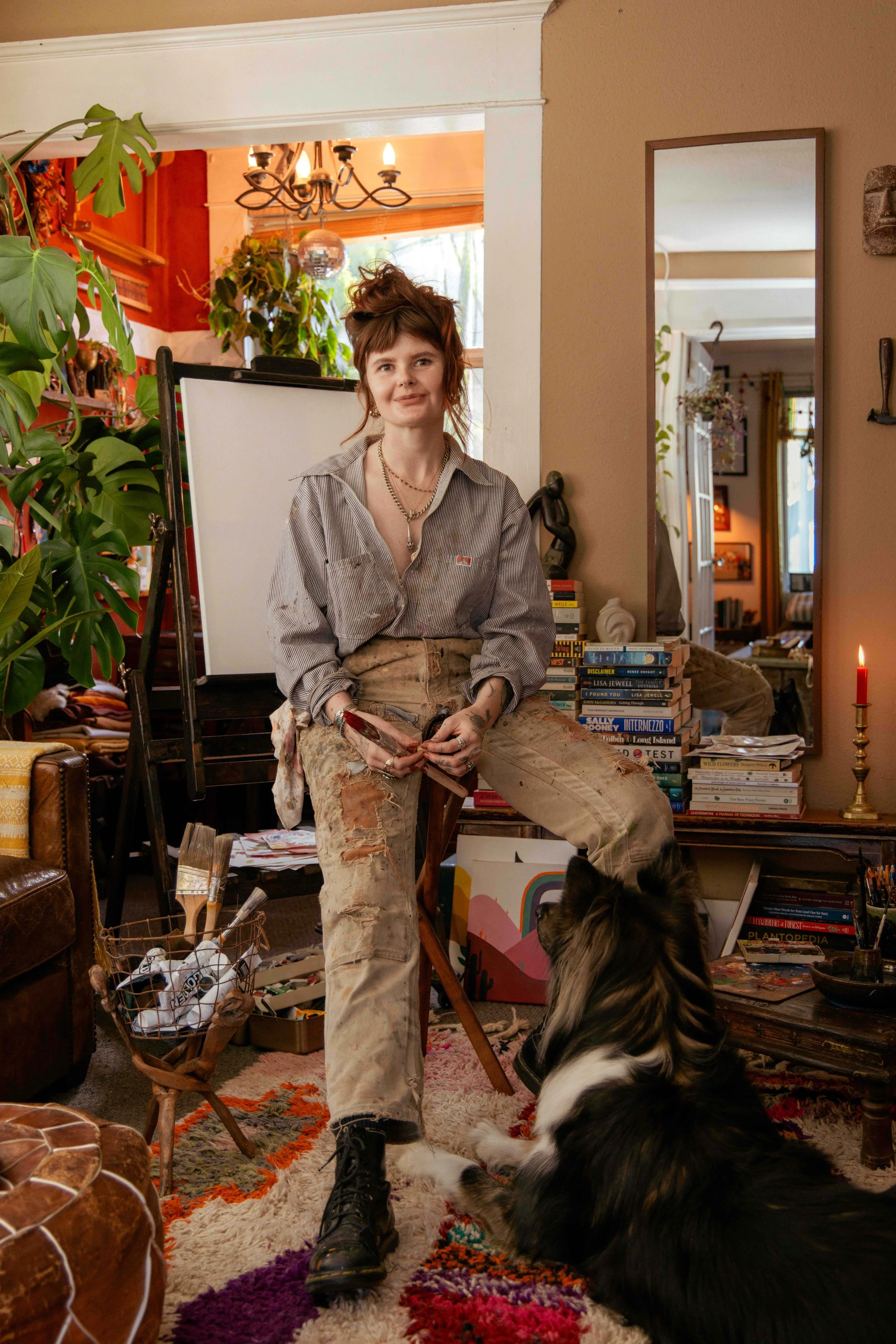 A woman with tattoos, messy hair, and casual clothes sitting in a bohemian-style living room with books, plants, and colorful decor, a large mirror, and a dog sitting at her feet.