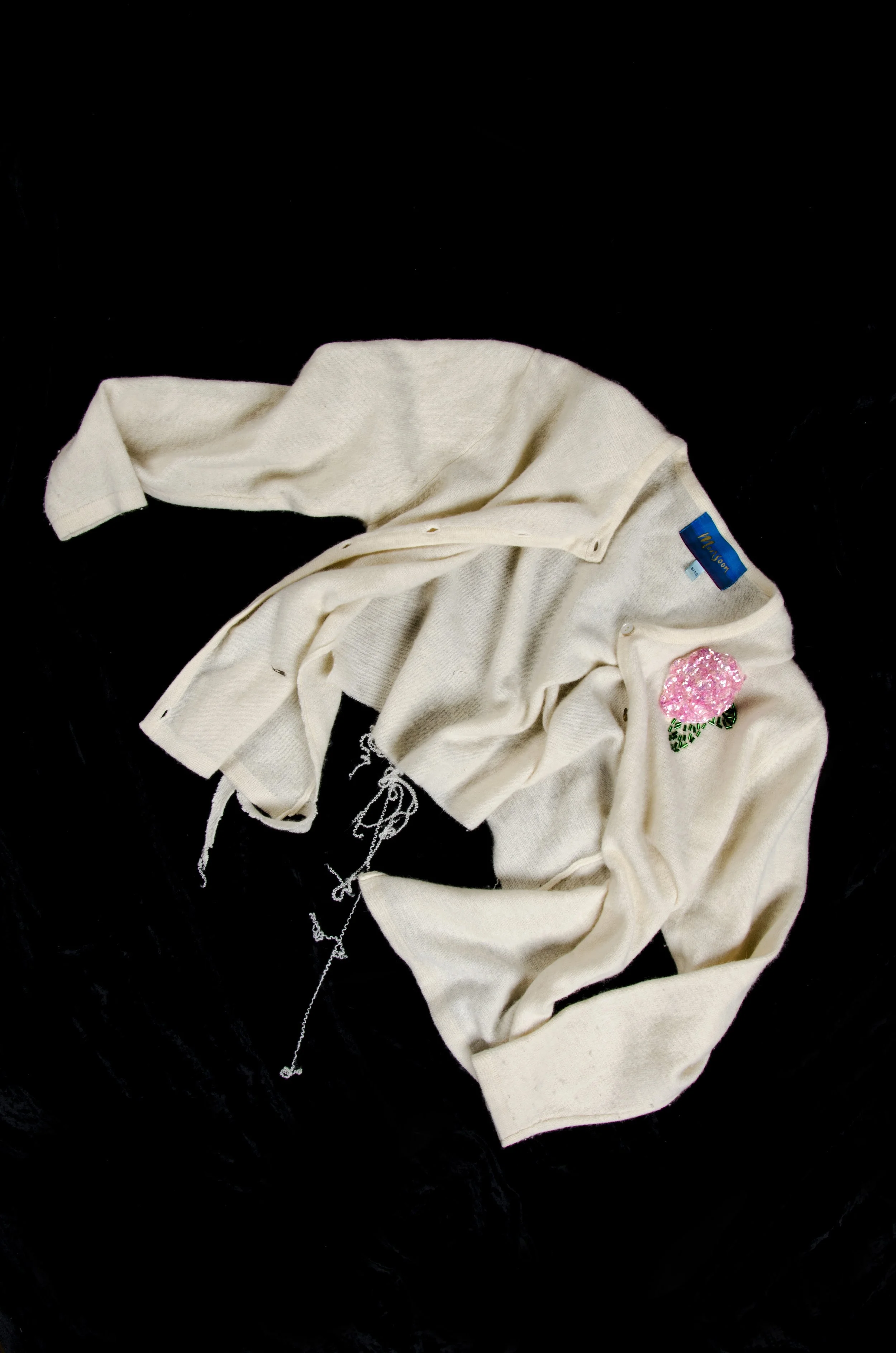 A crumpled off-white cardigan with a pink floral patch and a blue tag on a black background.