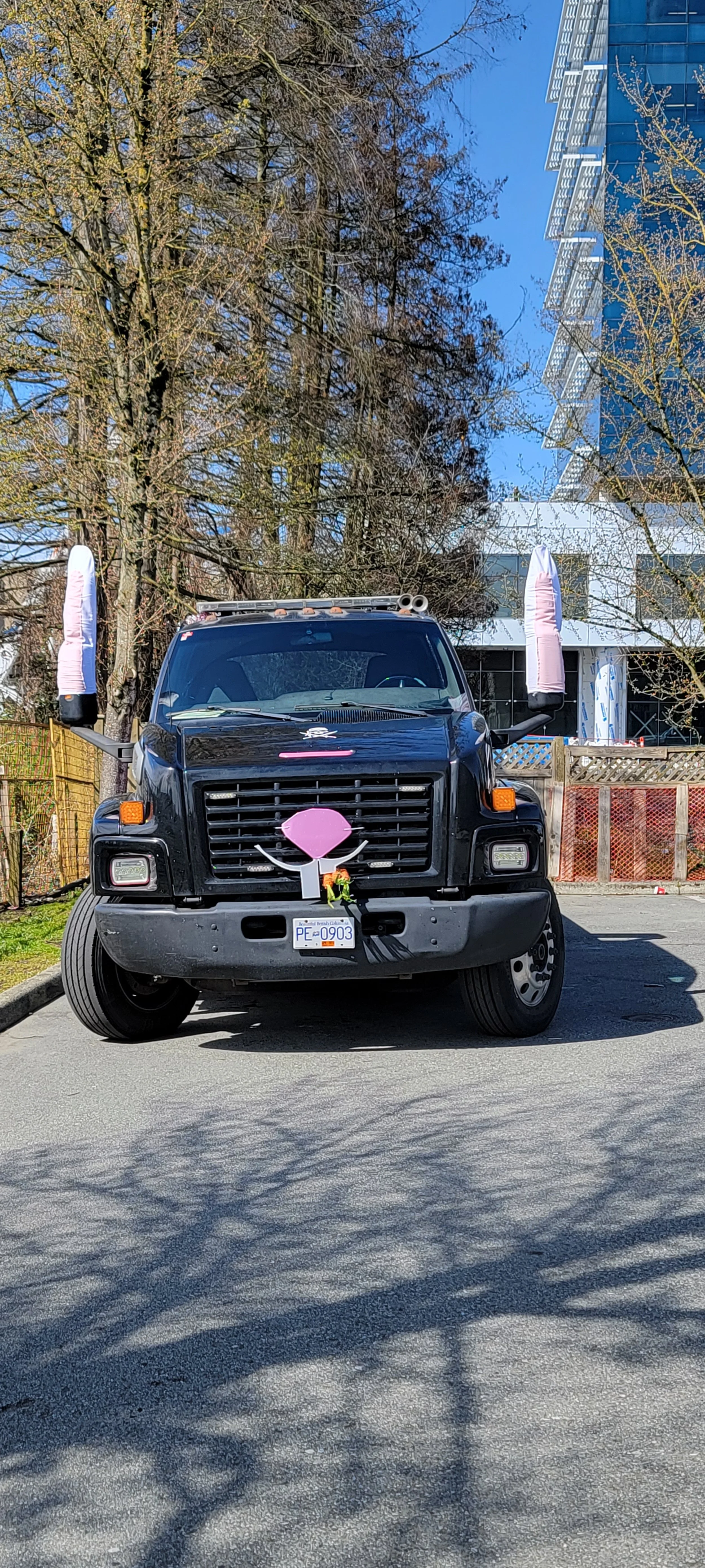 A black truck decorated with pink and white accents, including large sock-like objects on the side mirrors, large pink nose and closed eyes on the front grille, and a small pink bow on the hood, parked outdoors in front of trees and modern buildings.
