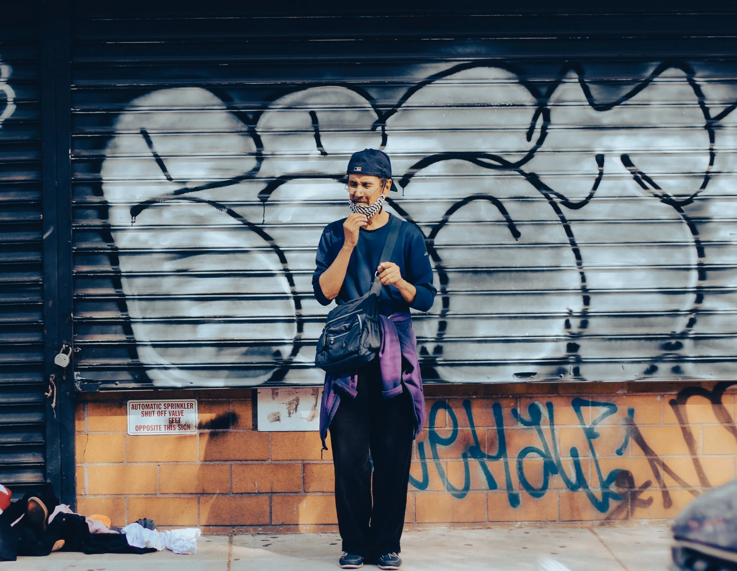 A young man in a dark blue shirt, black pants, and baseball cap, holding a purple jacket and wearing a face mask, standing in front of a graffiti-covered metal storefront with a brick base.