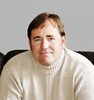 A man with short brown hair wearing a white sweater, sitting indoors with a neutral gray background.
