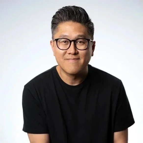 Portrait of a man with glasses and dark hair, wearing a black t-shirt, smiling slightly against a plain light background.