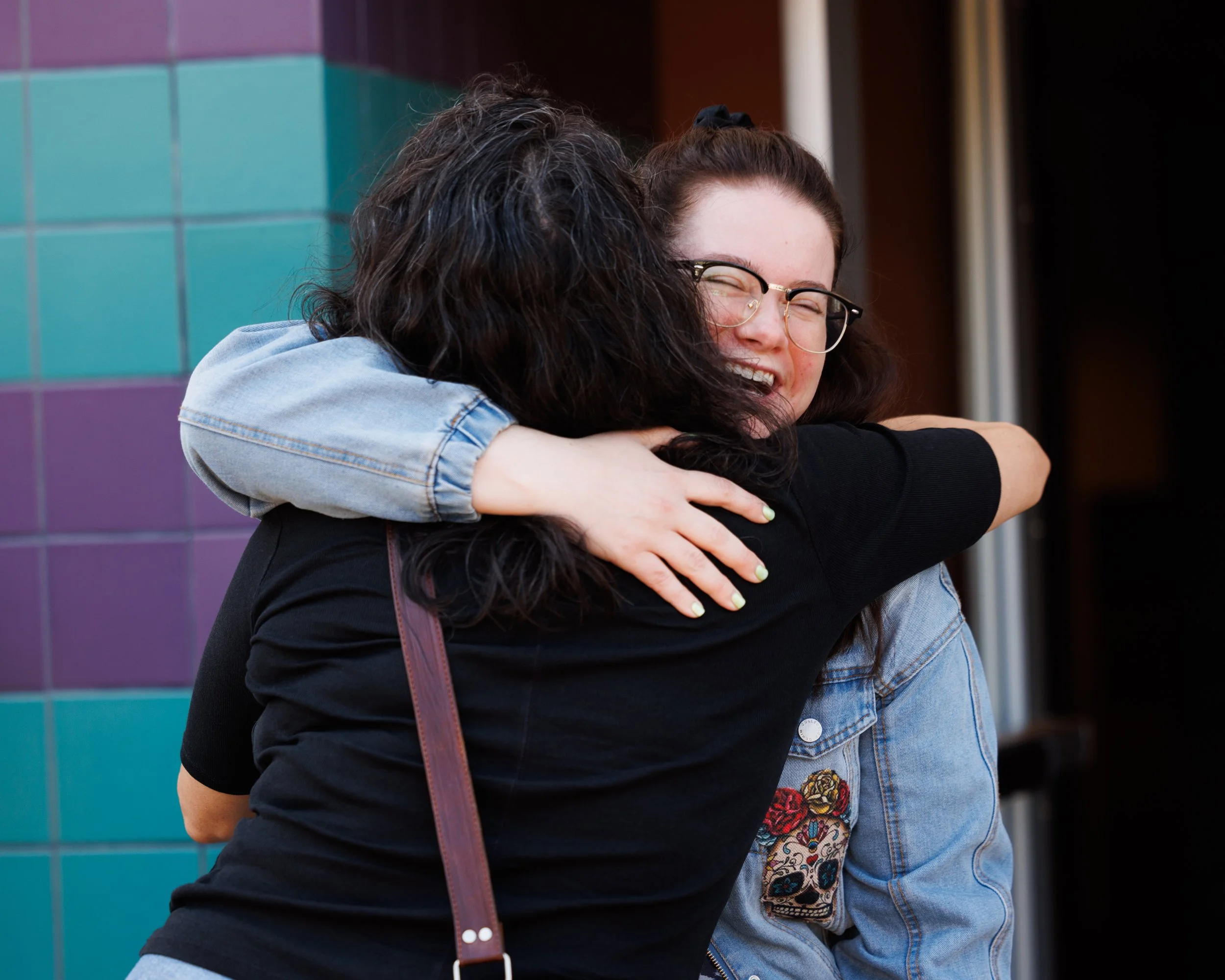 Two young women hugging each other and smiling, one with glasses and the other with dark curly hair, standing outdoors with colorful tiled wall in the background.