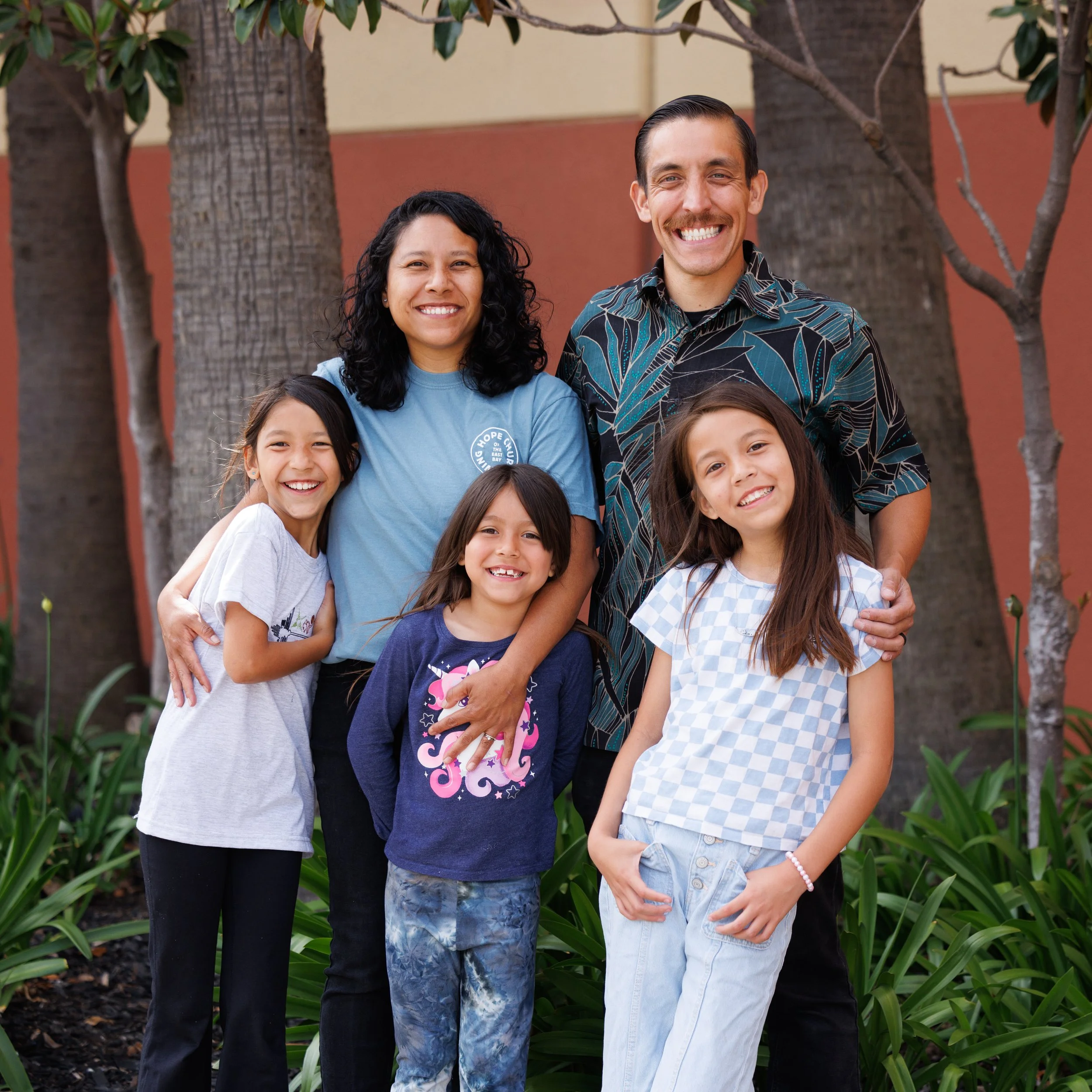 A diverse family of six smiling and posing outdoors in front of trees and a building wall.