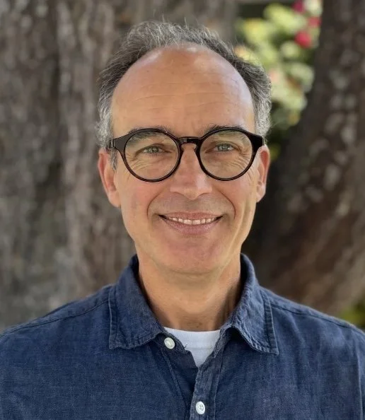 A middle-aged man with glasses smiling outdoors in front of a tree and flowers.