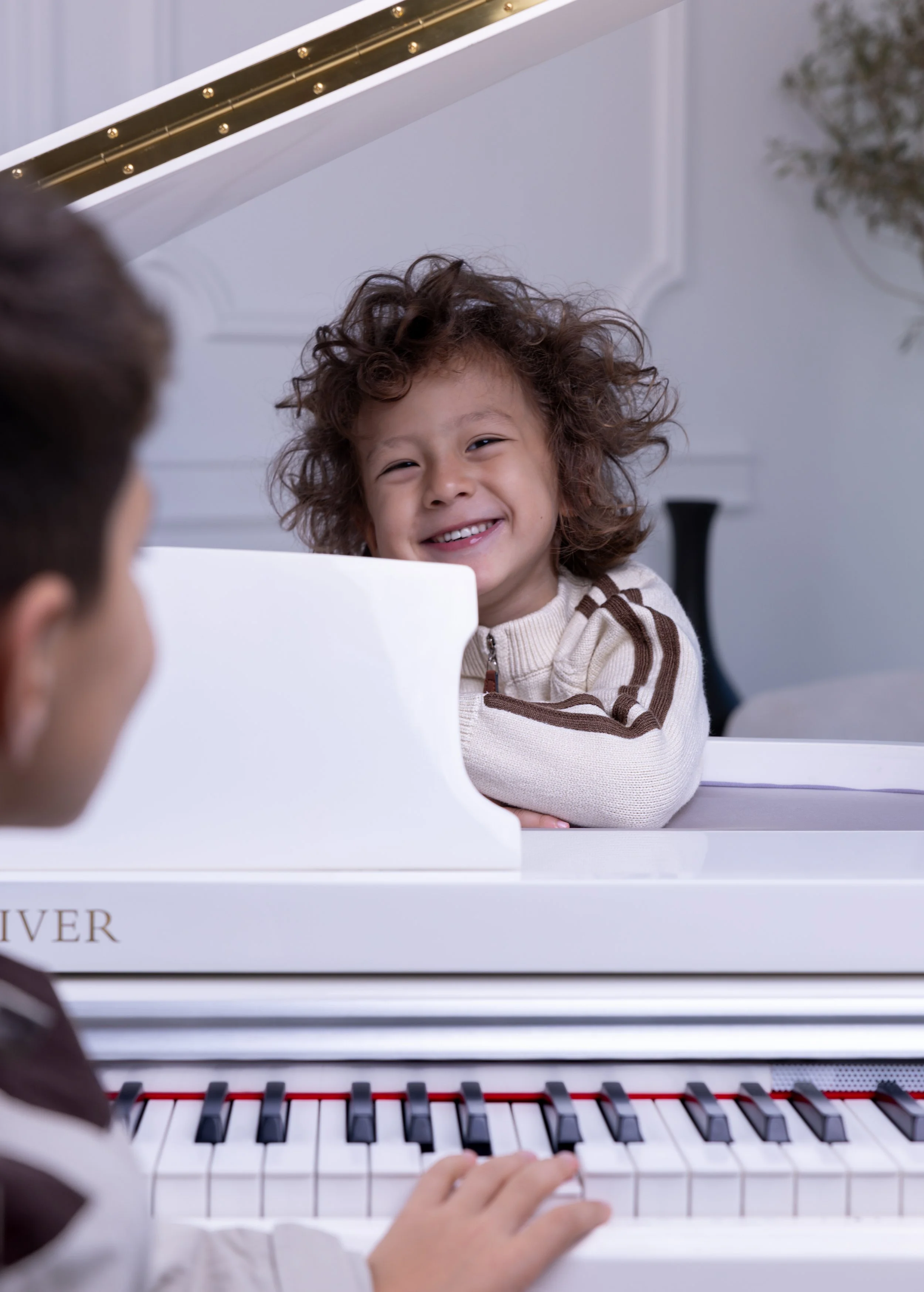 A young girl with curly hair smiling as she watches a person playing a white grand piano.