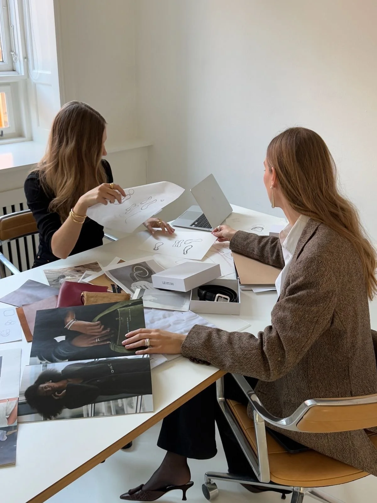 Two women sitting at a desk discussing fashion designs and looking at photos of models. The desk is cluttered with magazines, papers, a laptop, and a shoe box.