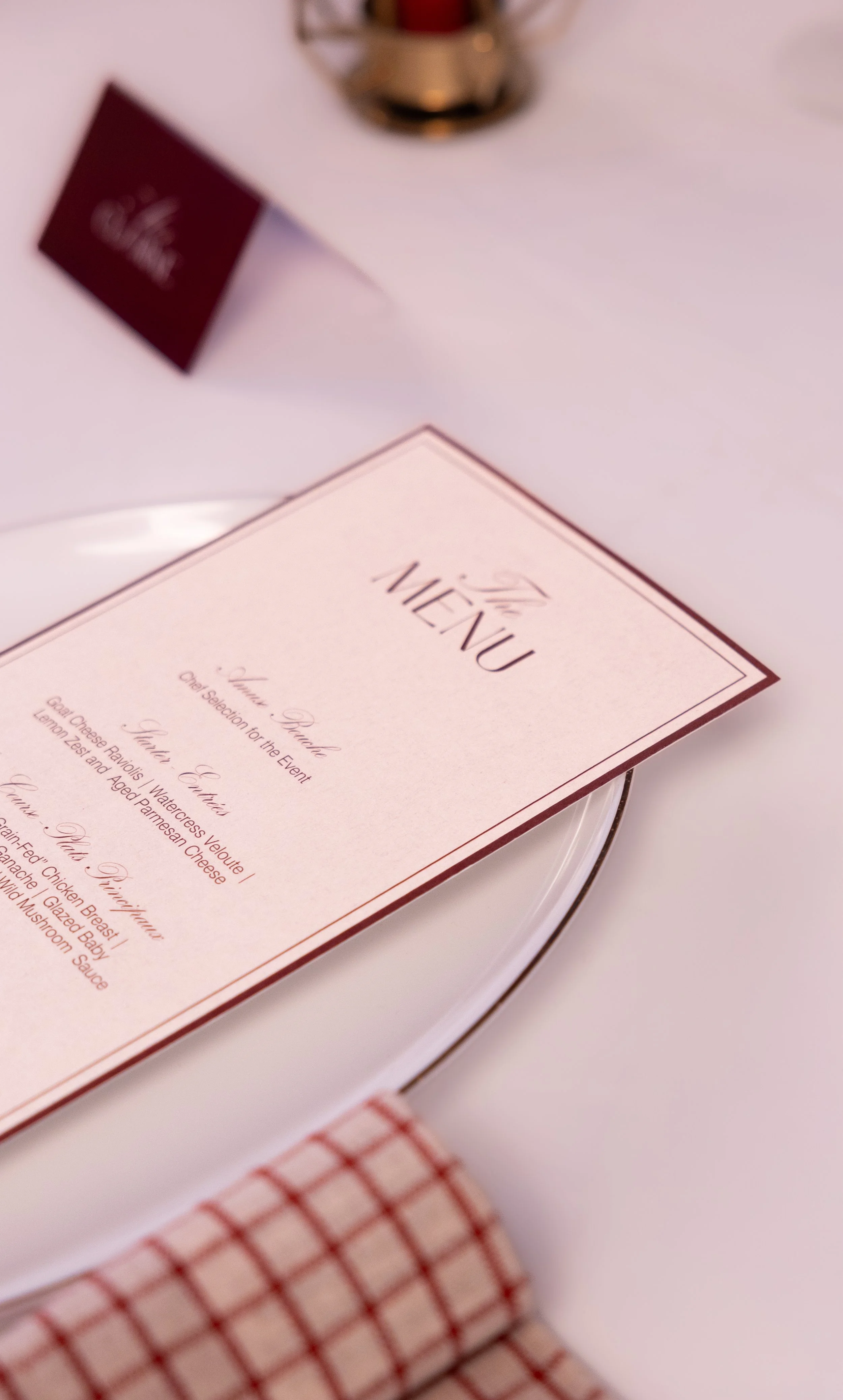Close-up of a menu card on a white table, with a red and white checkered napkin and a blurred red card and a gold object in the background.