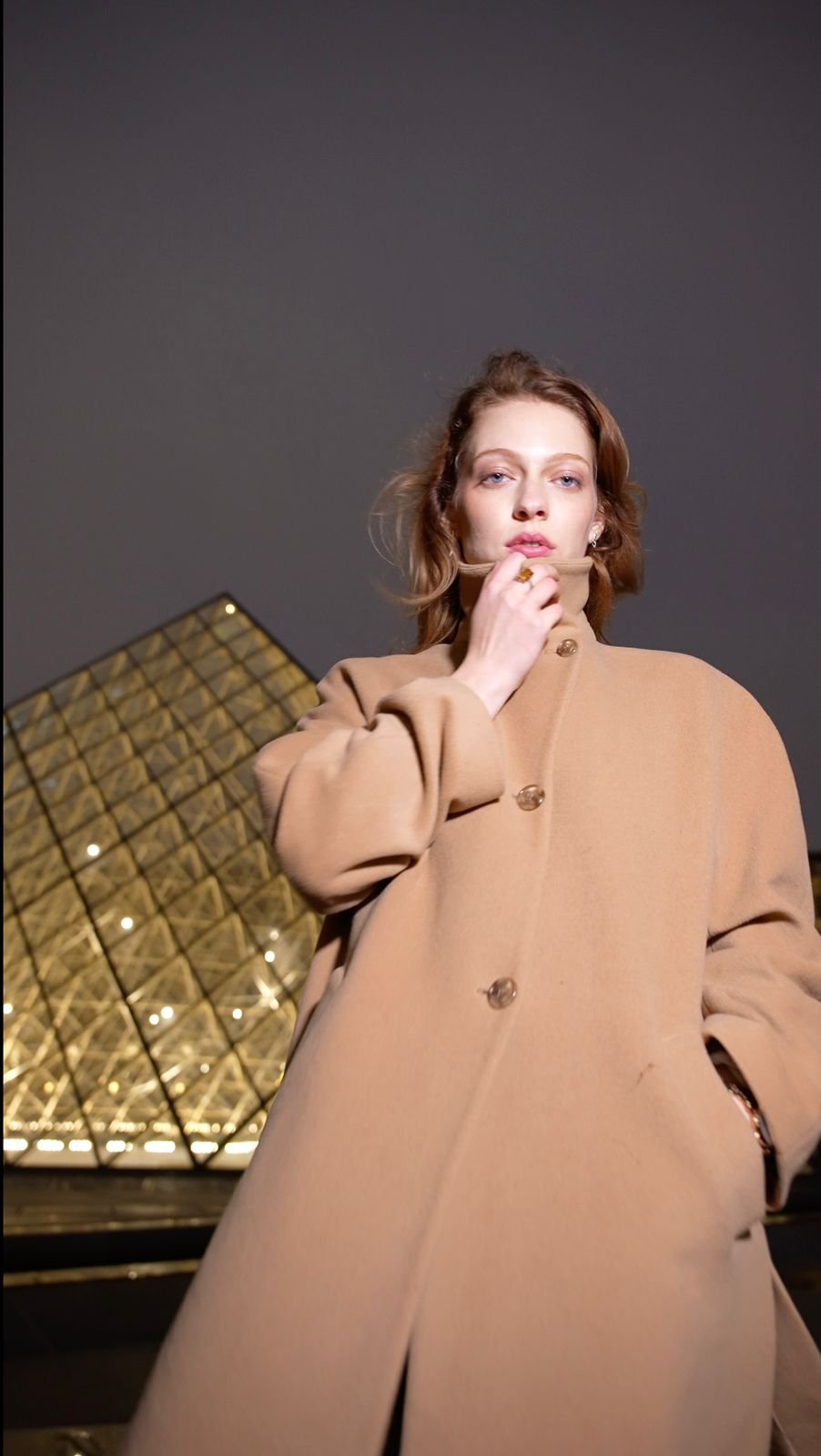 A young woman with wavy brown hair and blue eyes wearing a tan coat with large buttons, standing outdoors in front of a modern glass building at night.